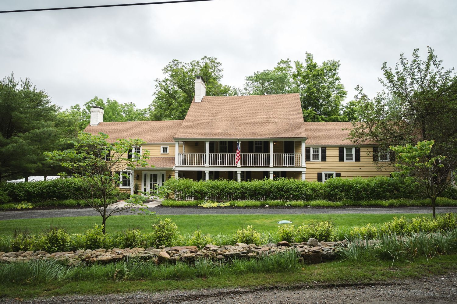 A large beige colonial style house with wraparound porch sits nestled among landscaped gardens and trees.
