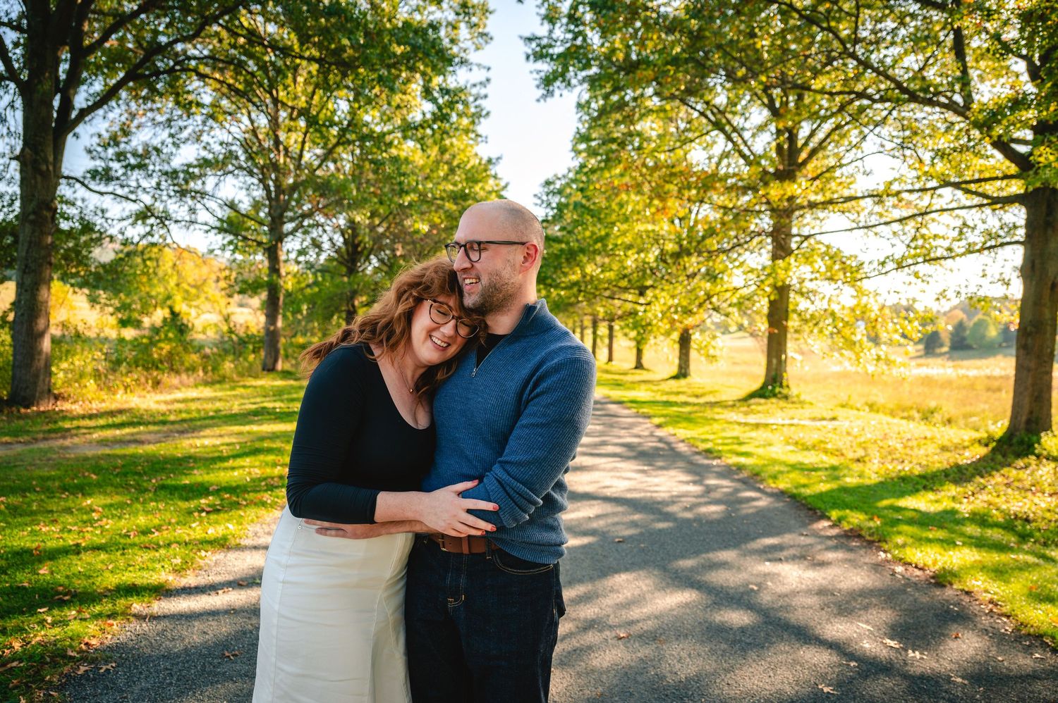 A couple embraces on a sunlit path surrounded by golden autumn trees in a park setting.