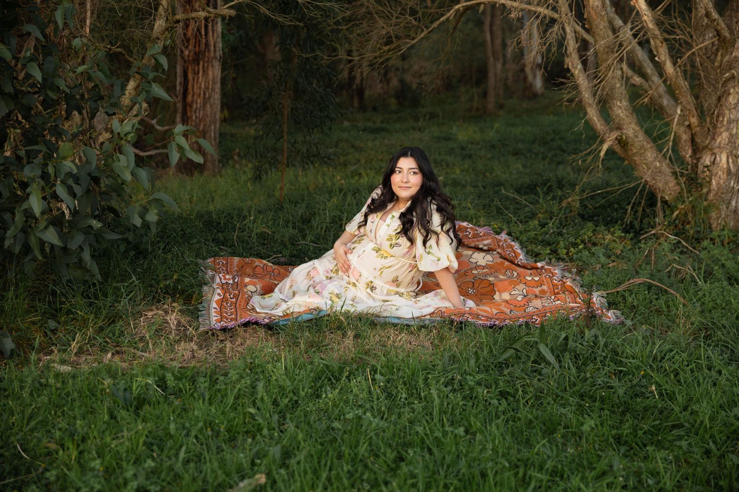 A maternity portrait on a vintage blanket in a wooded outdoor setting.