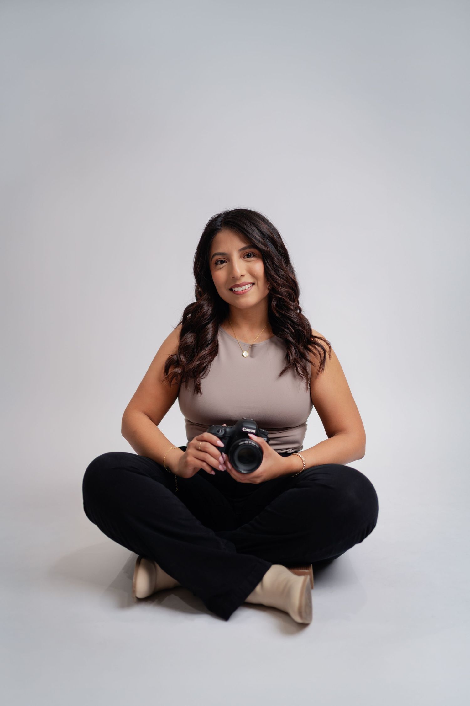 Female photographer sitting cross-legged on floor holding DSLR camera, smiling warmly in a studio setting.