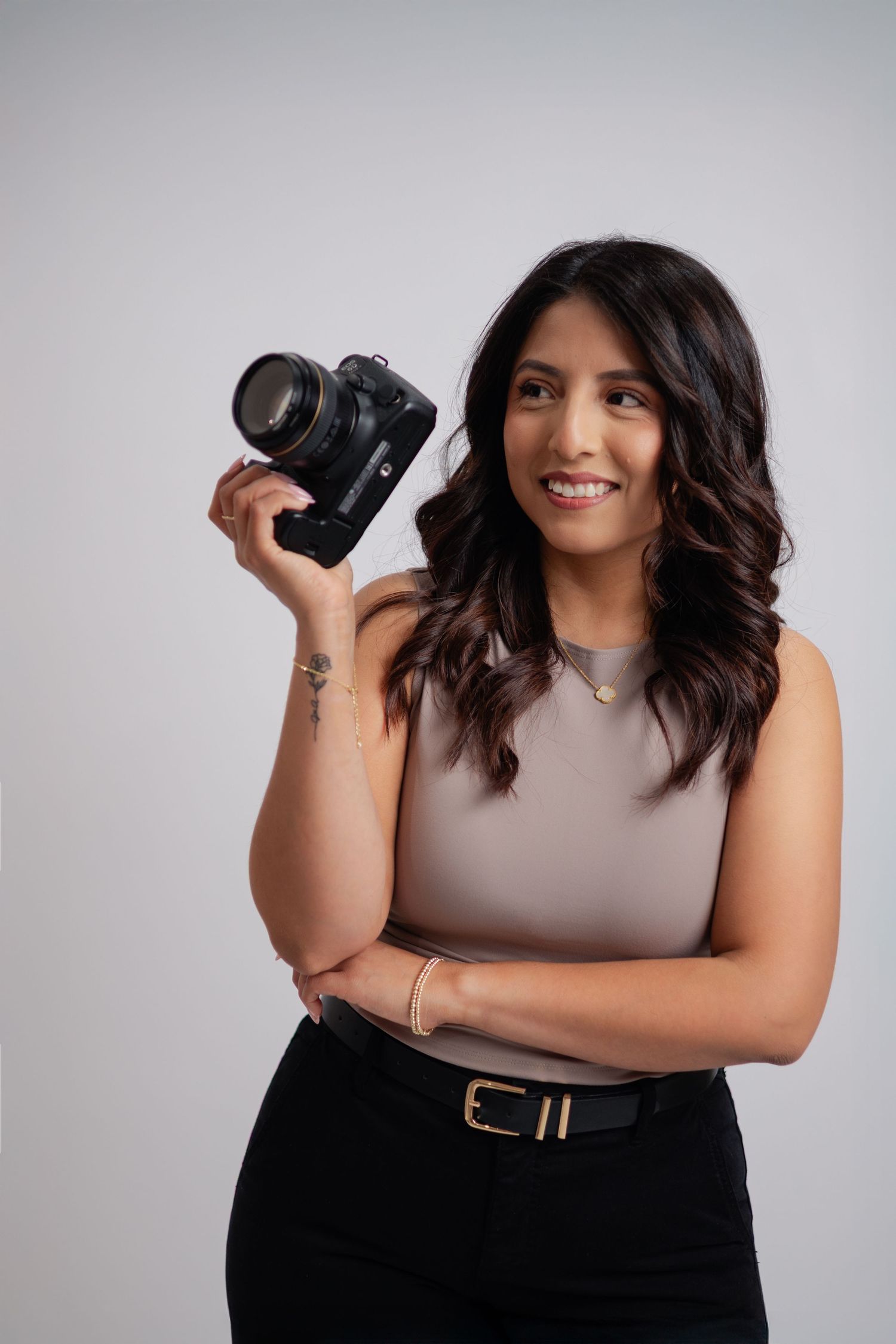 A smiling female photographer holds a DSLR camera while posing in a beige sleeveless top and black pants on gray background.