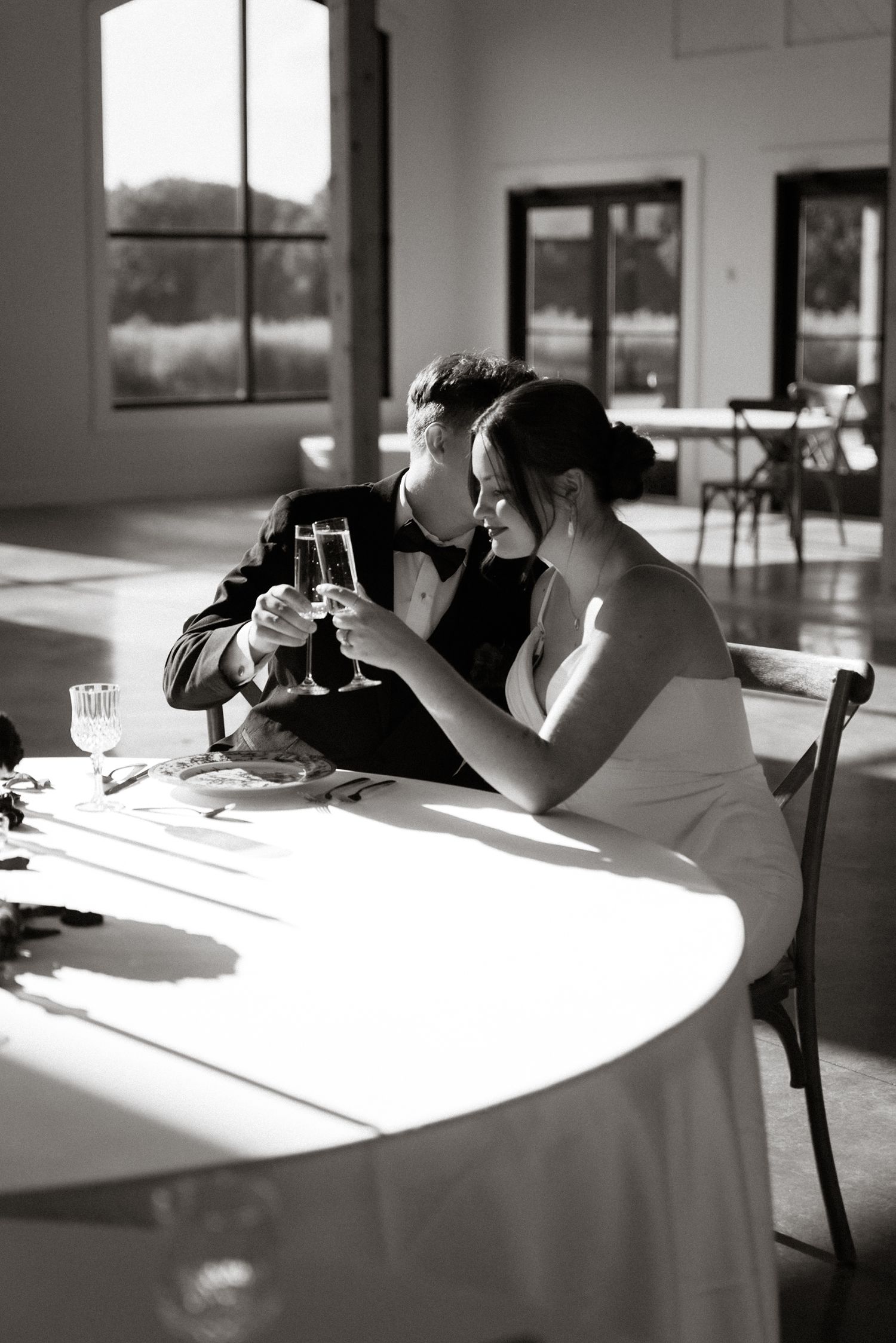 A black and white wedding photo showing a couple toasting with champagne glasses in a sunlit room.