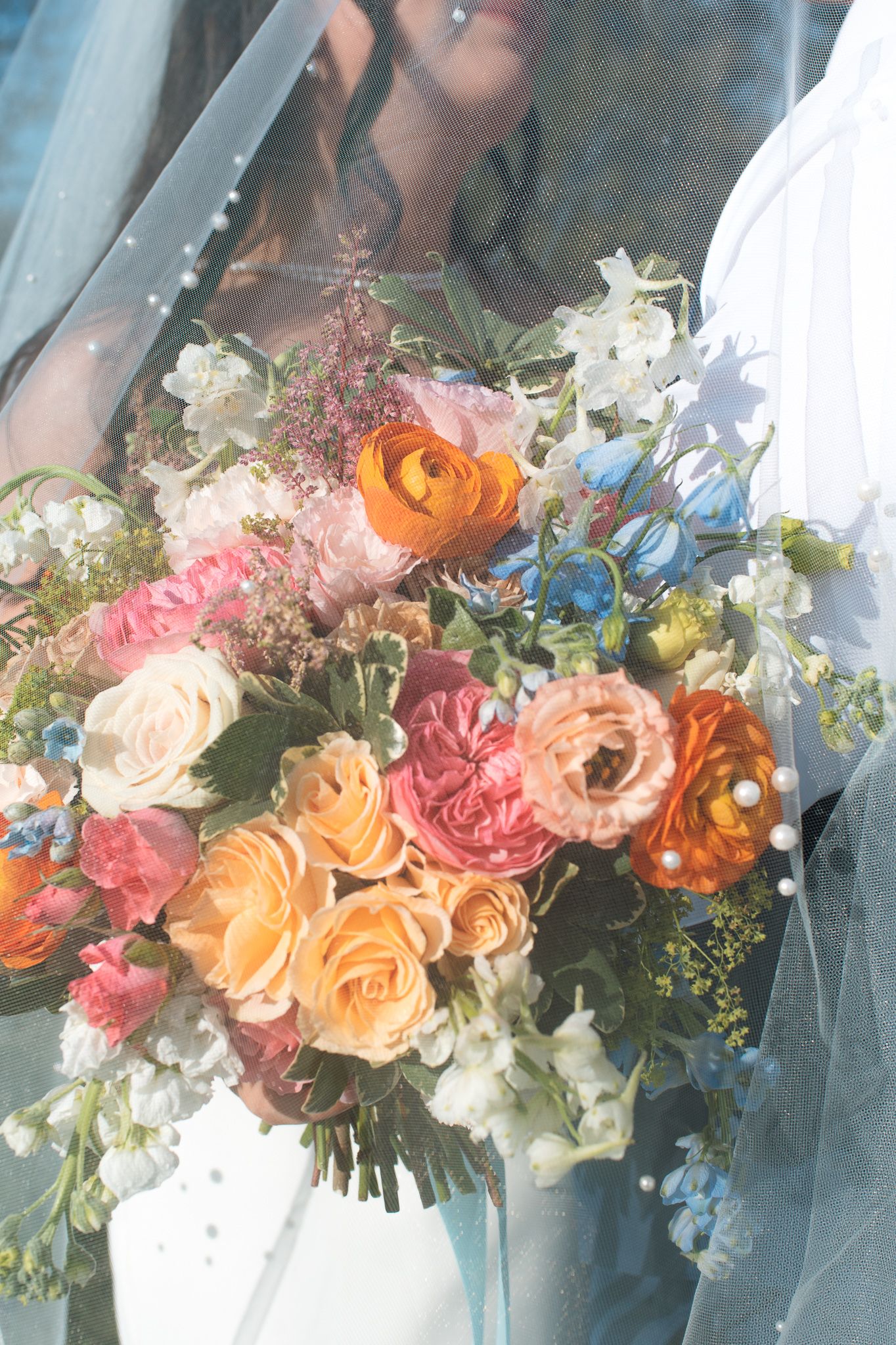 Bride holding a vibrant wedding bouquet with peach roses, pink ranunculus, and blue flowers under a flowing veil.