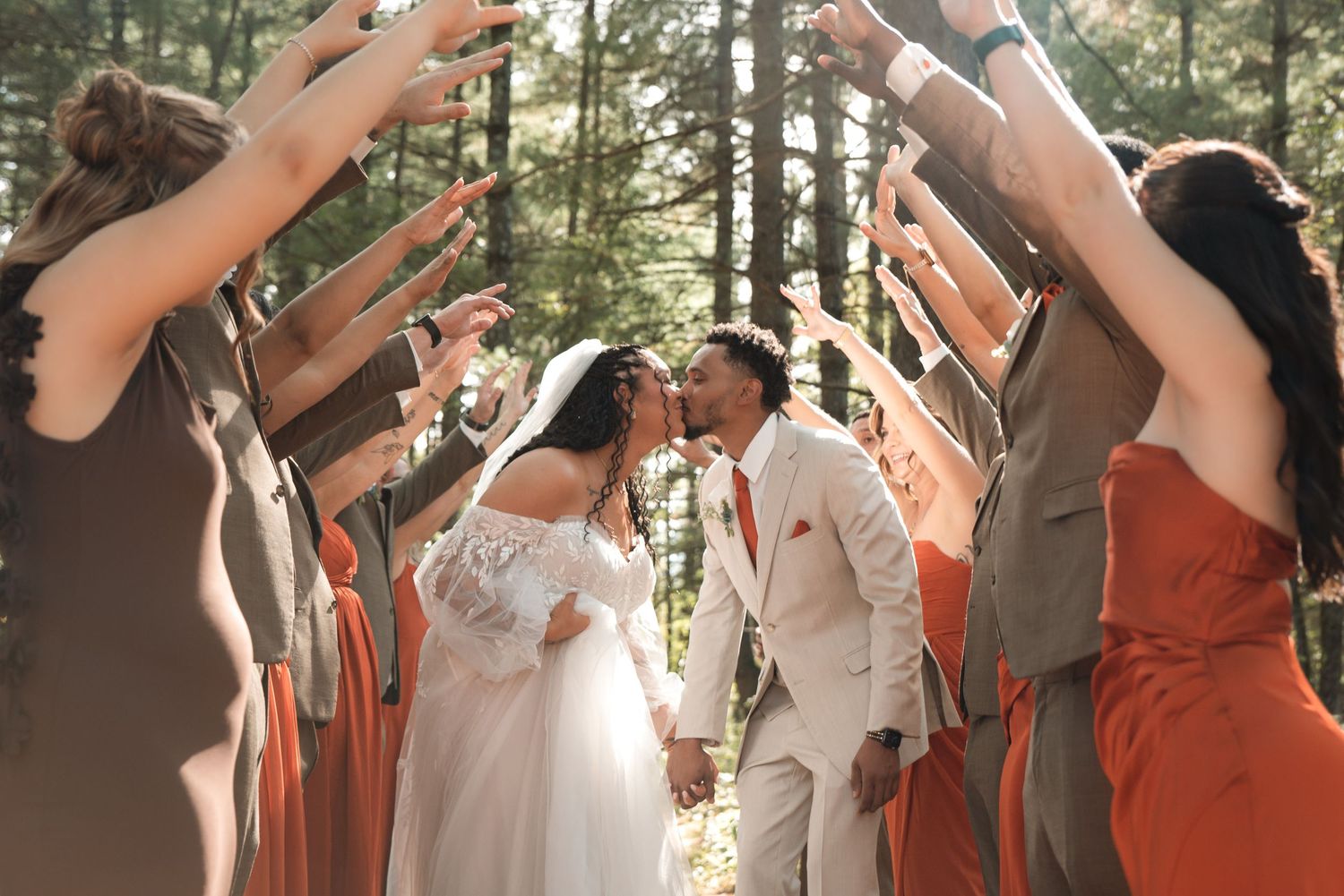 Bride and groom share a kiss in a sunlit forest as bridesmaids and groomsmen raise their arms to form an arch around the couple.