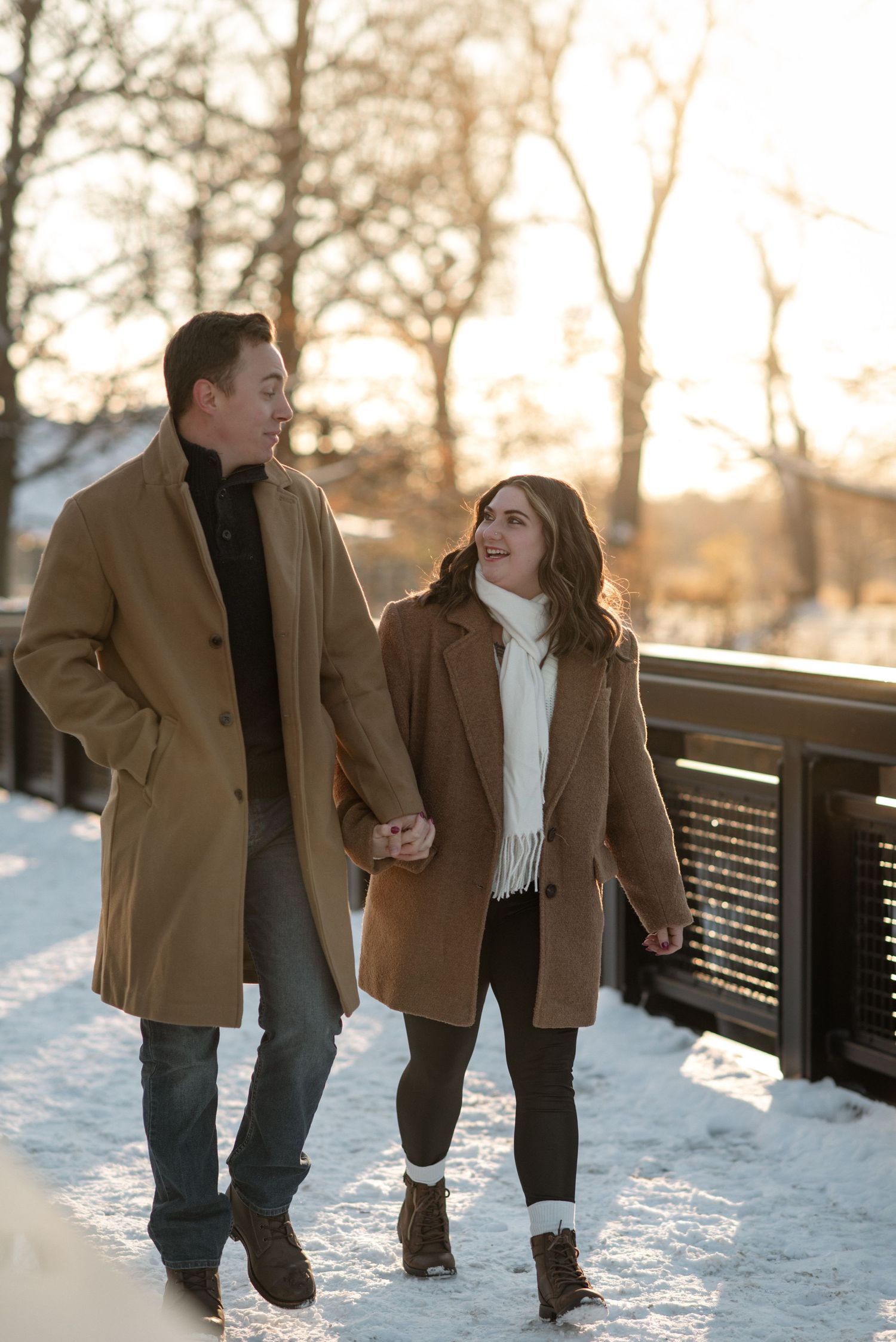 A couple in matching tan coats walking together through snowy scenery at sunset.