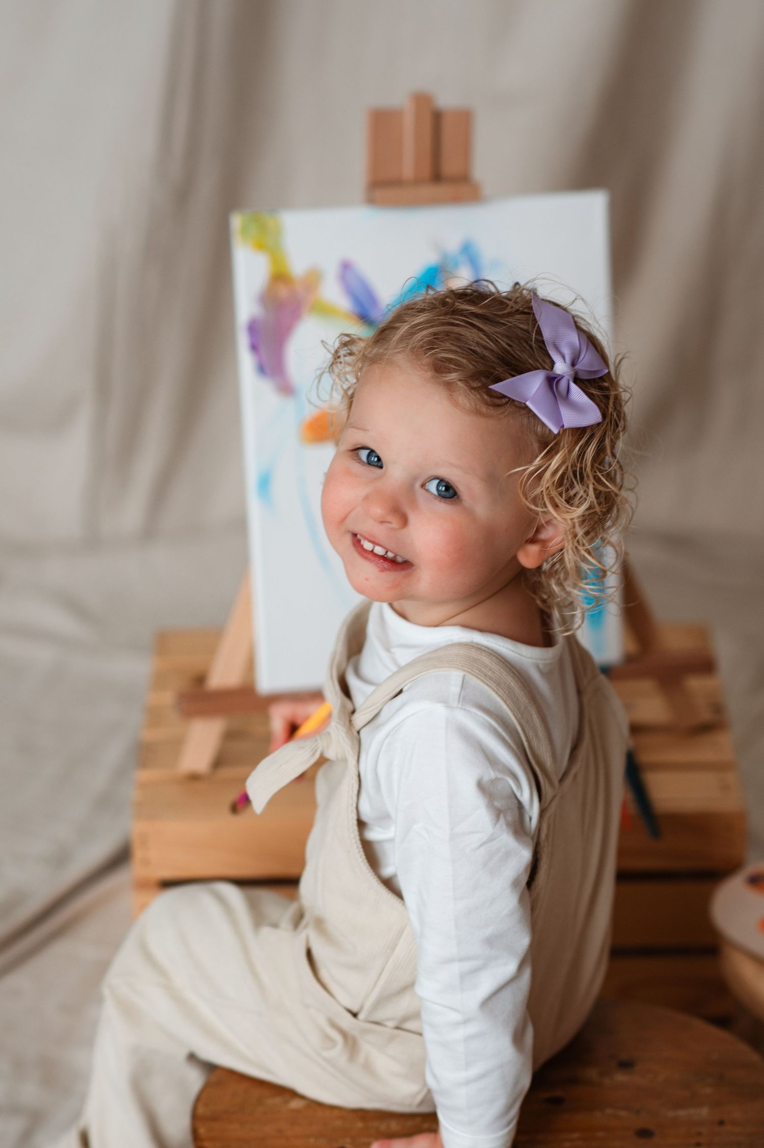 A small child wearing white clothing lies on their stomach while creating artwork at a wooden easel.