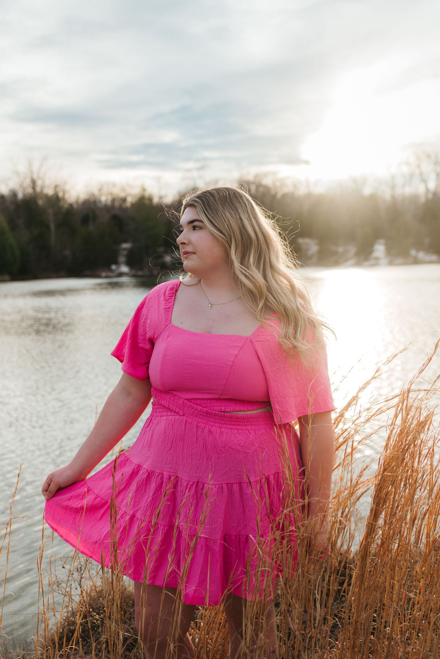 A person in a bright pink dress stands among tall golden grass near a lake at sunset.