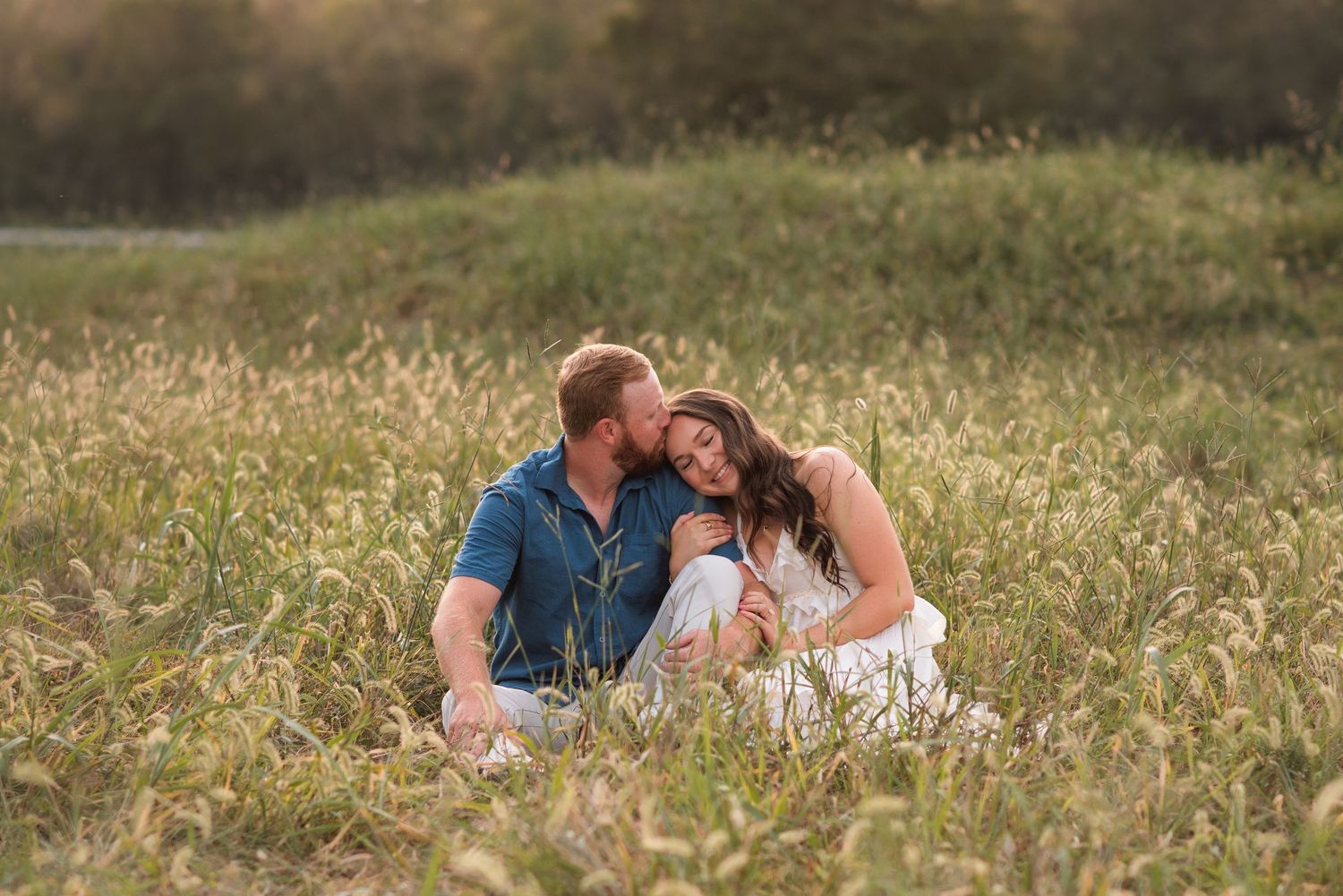 Couple sitting close together in a tall grassy field at golden hour, the woman in a white dress leaning against the man in a blue shirt.
