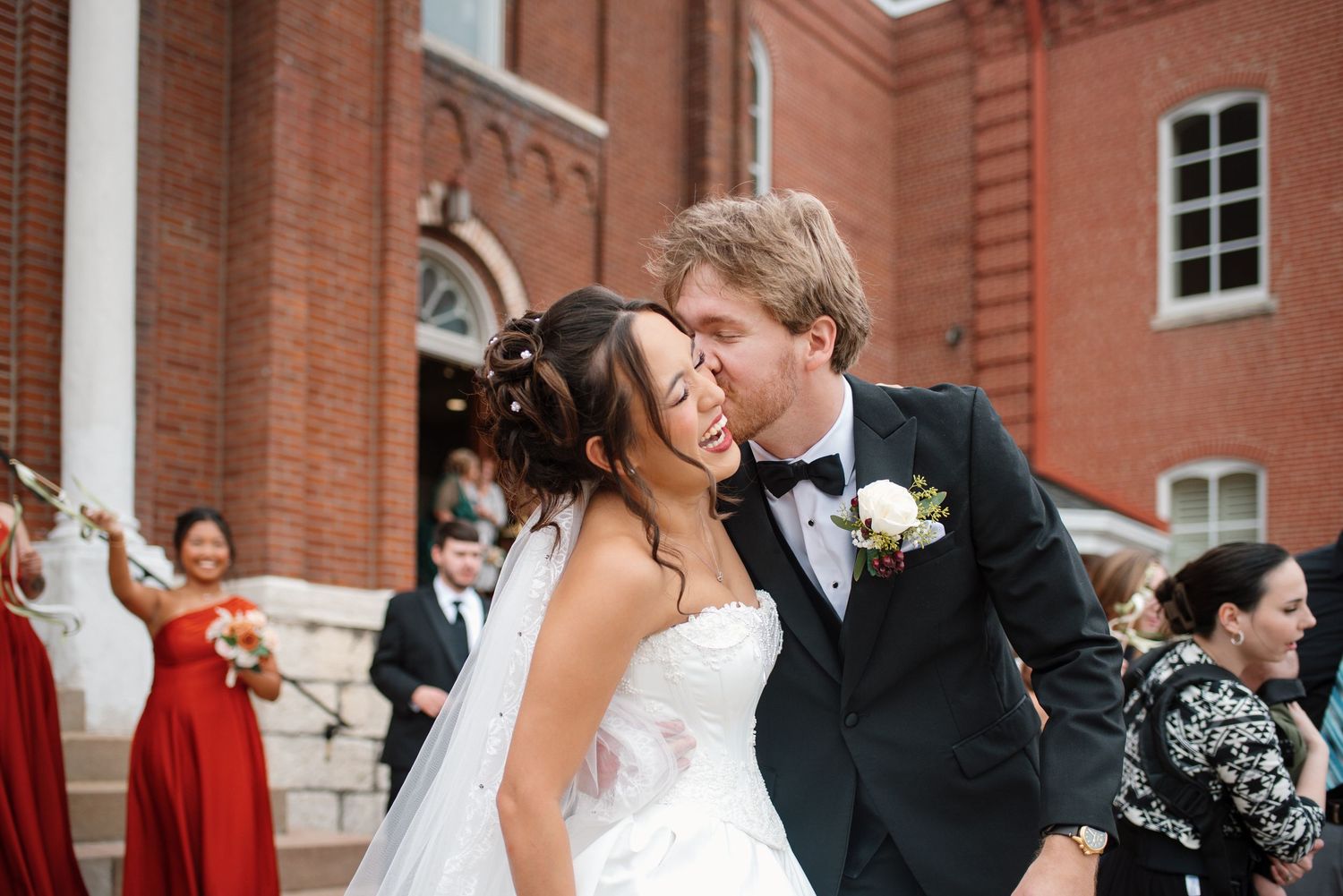 Groom in a black tuxedo kisses a laughing bride in a white wedding gown outside a red brick church as guests in red dresses celebrate.