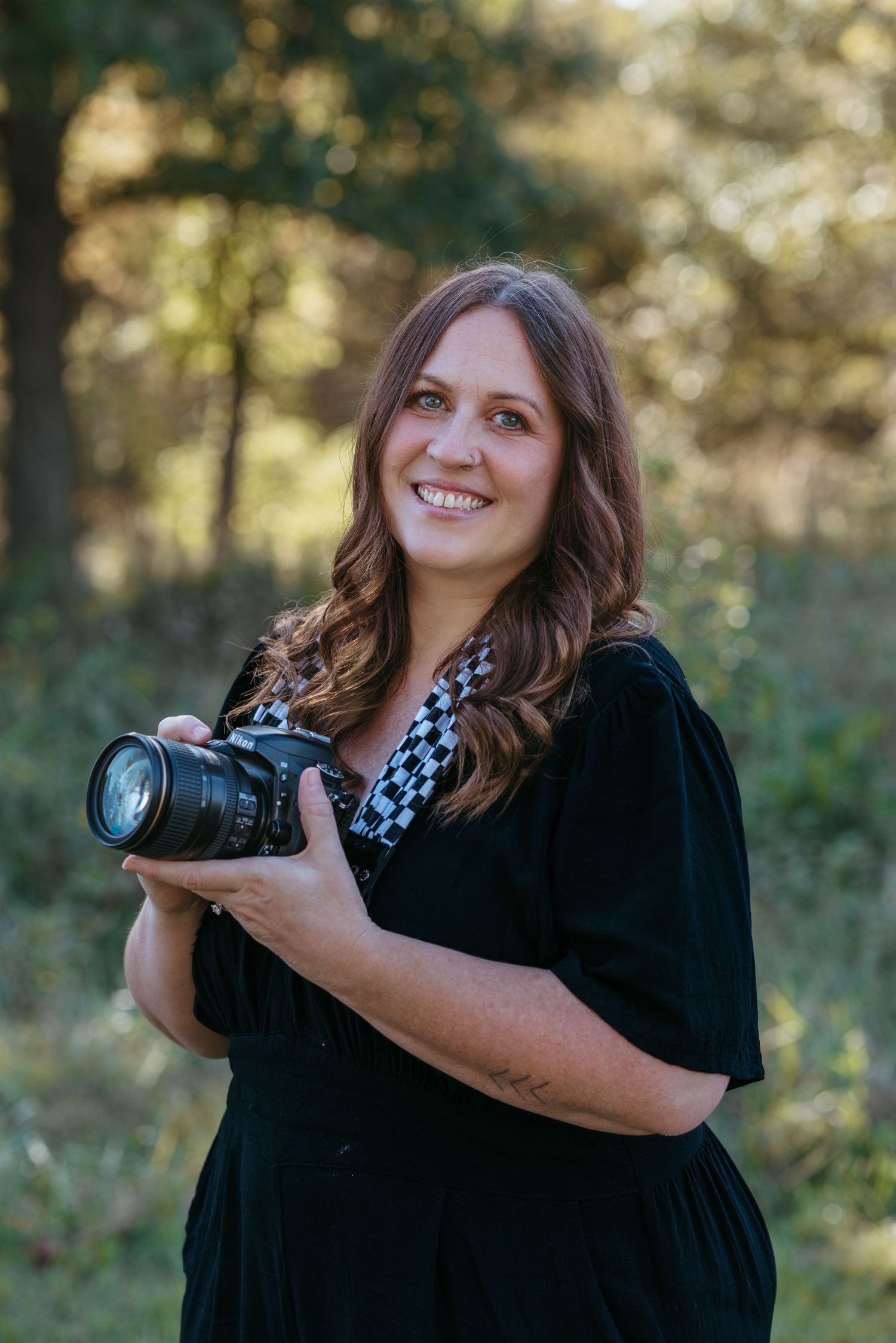 Professional photographer wearing black dress with checkered camera strap poses with DSLR camera in outdoor setting.
