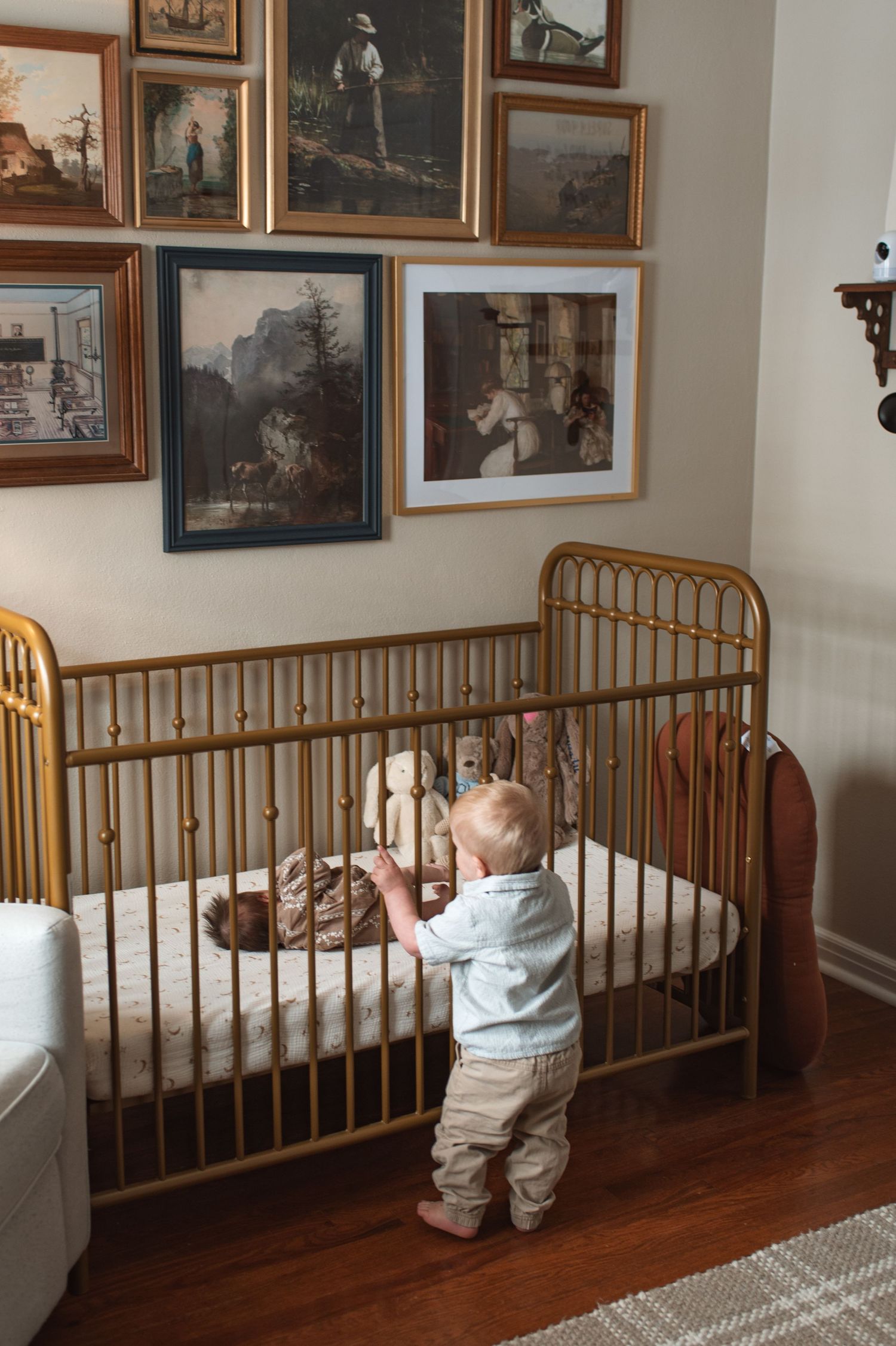 Baby in light colored clothing stands at wooden railing looking at framed artwork on gallery wall.