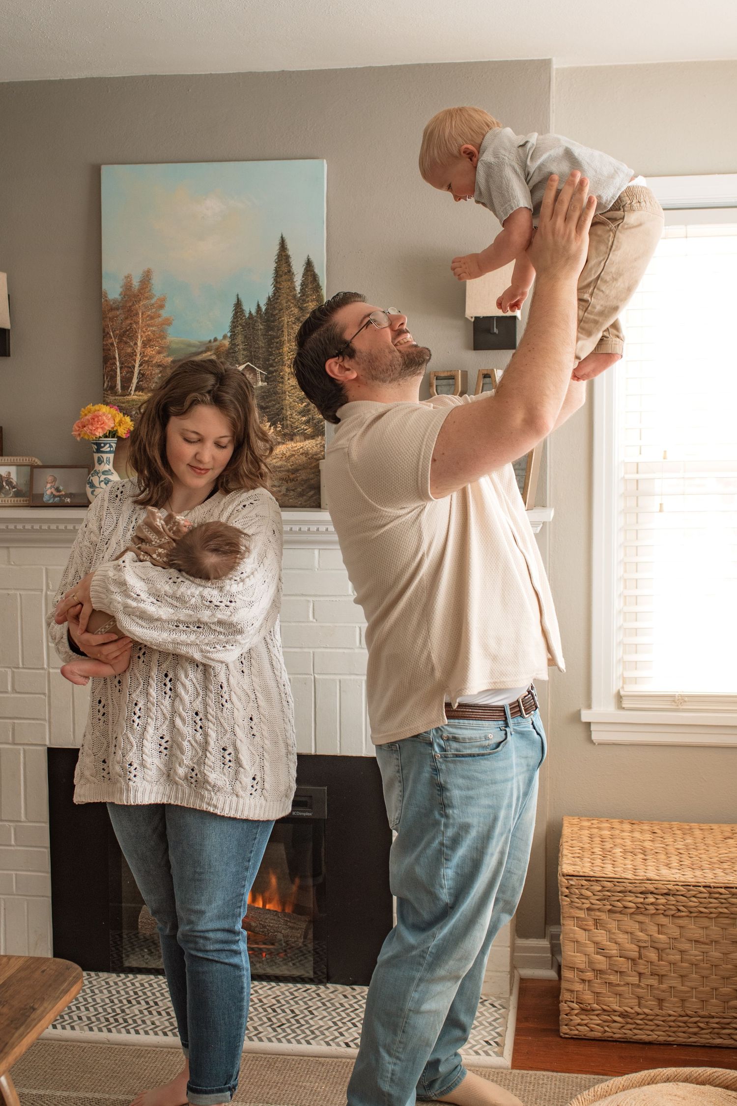 A couple in a cozy living room with a fireplace holds their young children in matching sweaters and jeans.