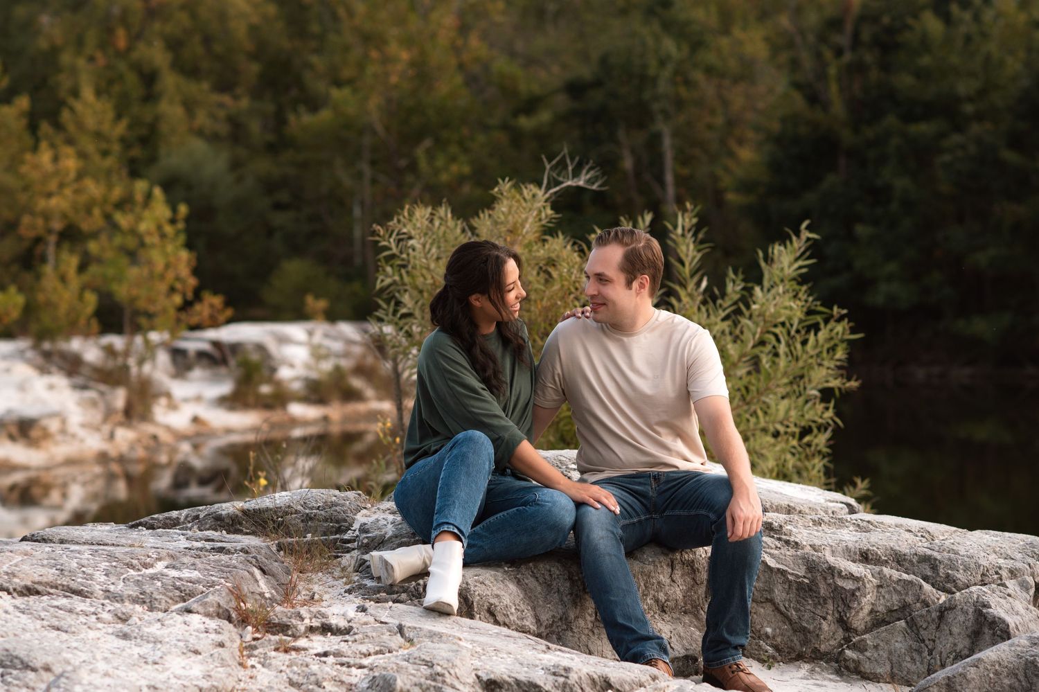 Couple sitting on rocky outcrop in nature, sharing a romantic moment with lush green trees in the background.