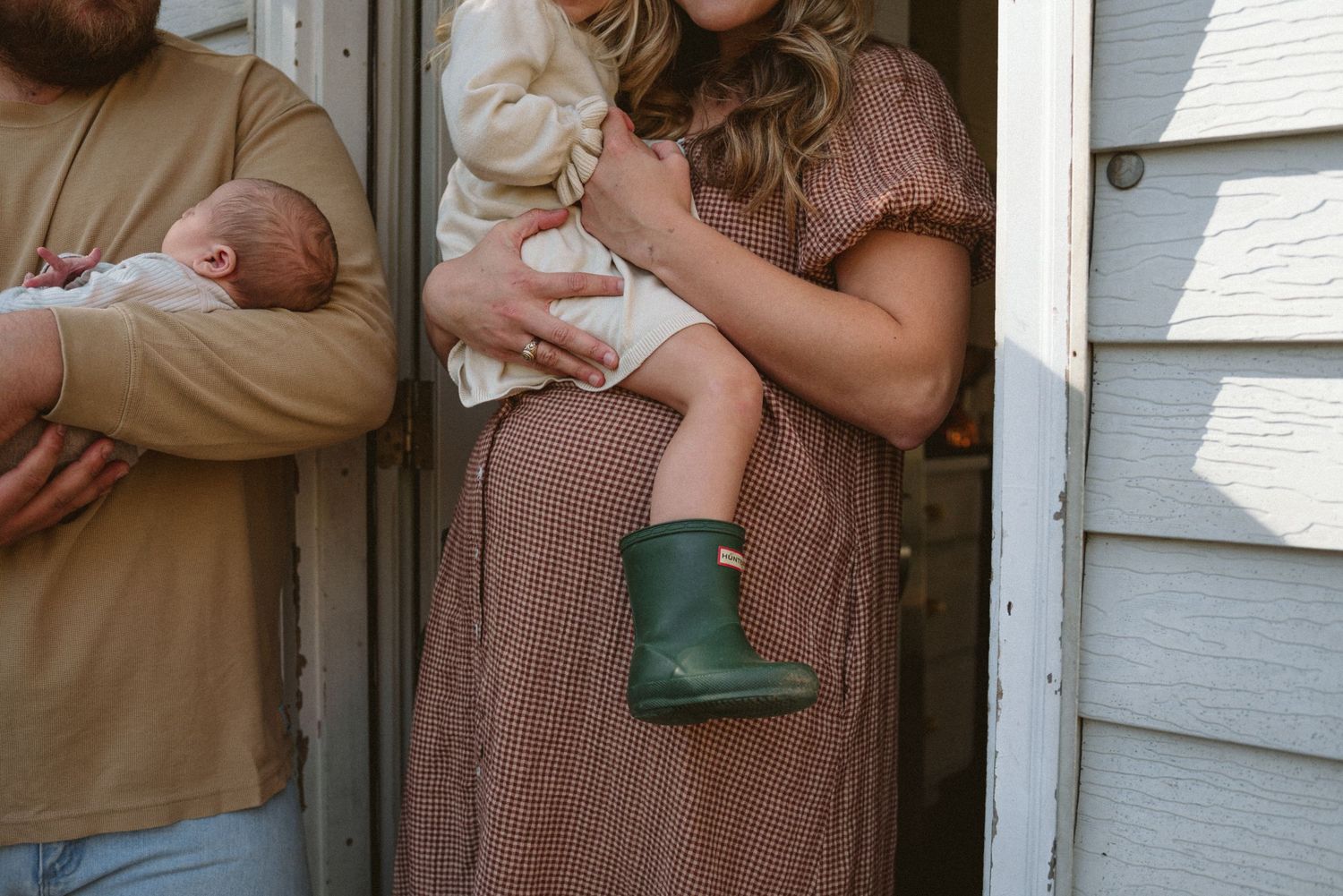 A mother in a brown dress holds a toddler in green rain boots while a man cradles a newborn outside a home.