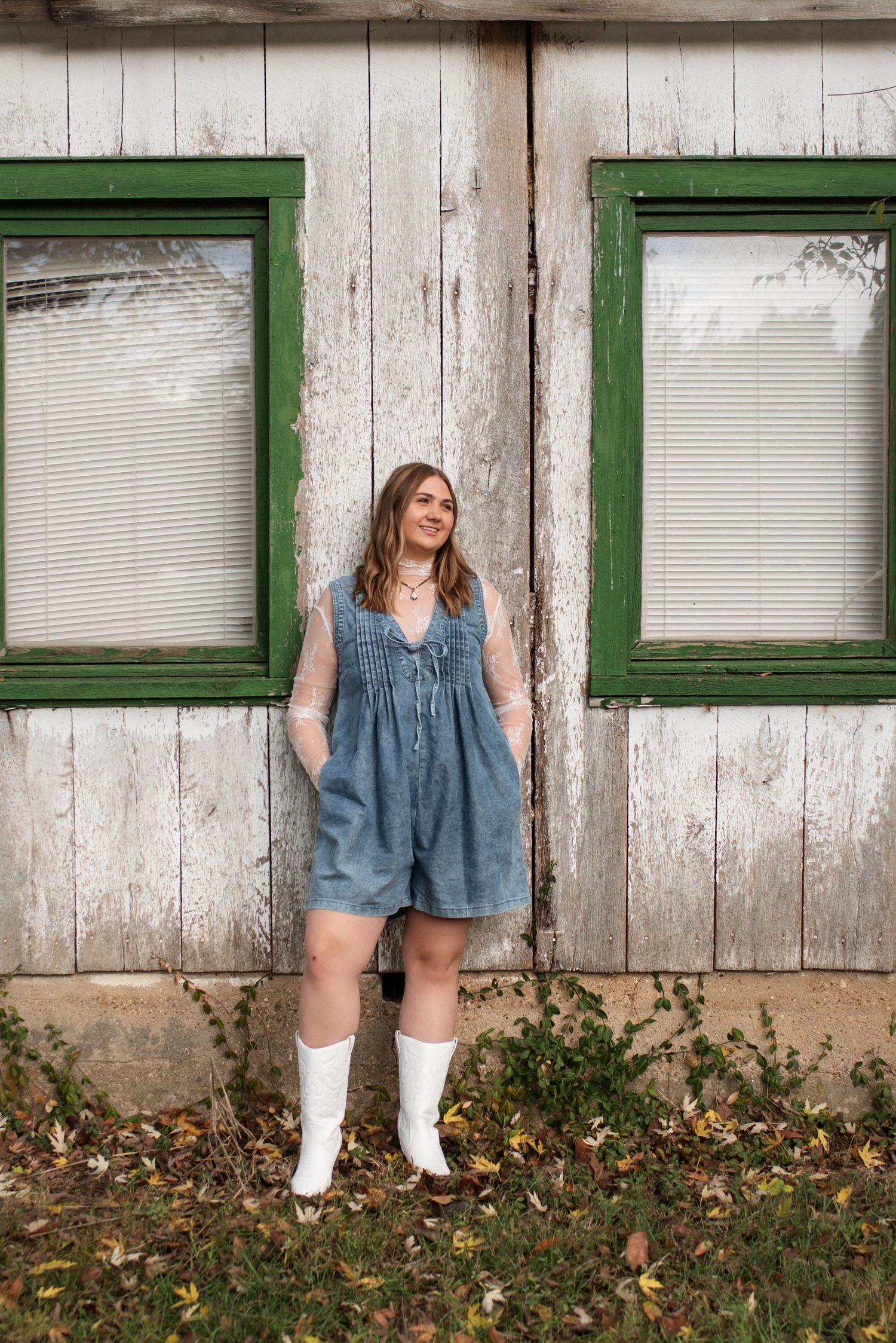 A denim dress and white boots outfit photographed against a rustic white wall with green-trimmed windows.