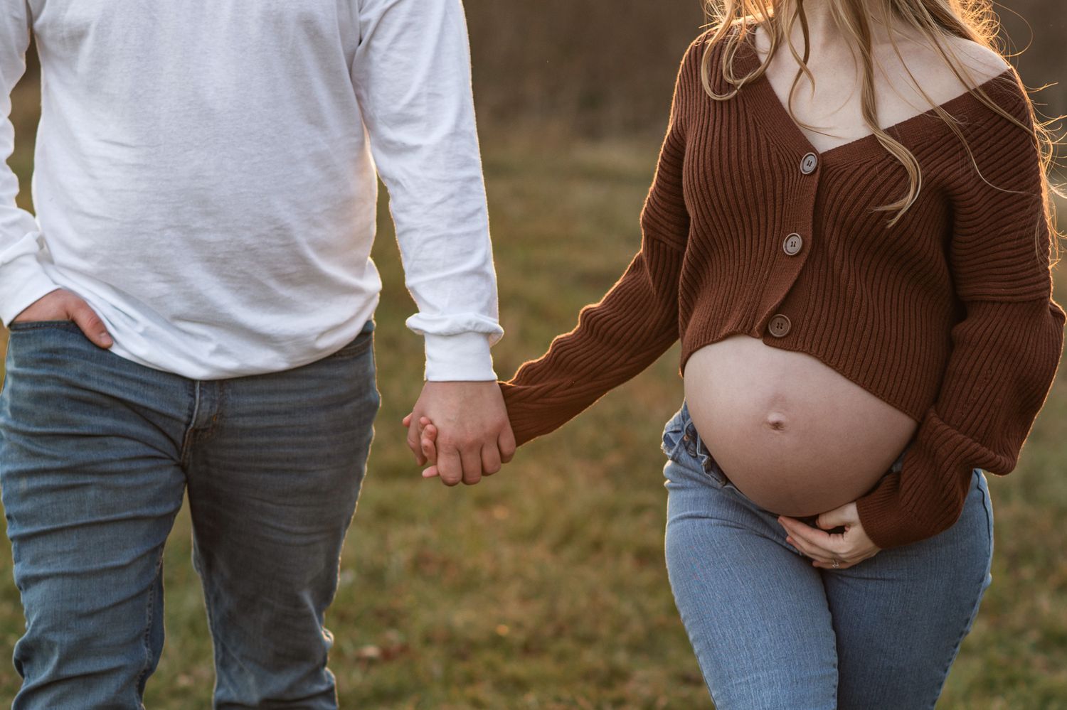 Pregnant woman in brown sweater and jeans holds partner's hand outdoors, cradling baby bump in a maternity photo.