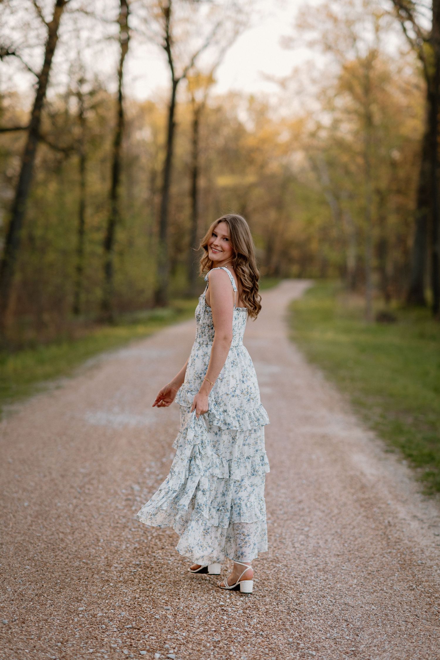 A child in a floral dress twirls and dances along a gravel path surrounded by autumn trees at sunset.
