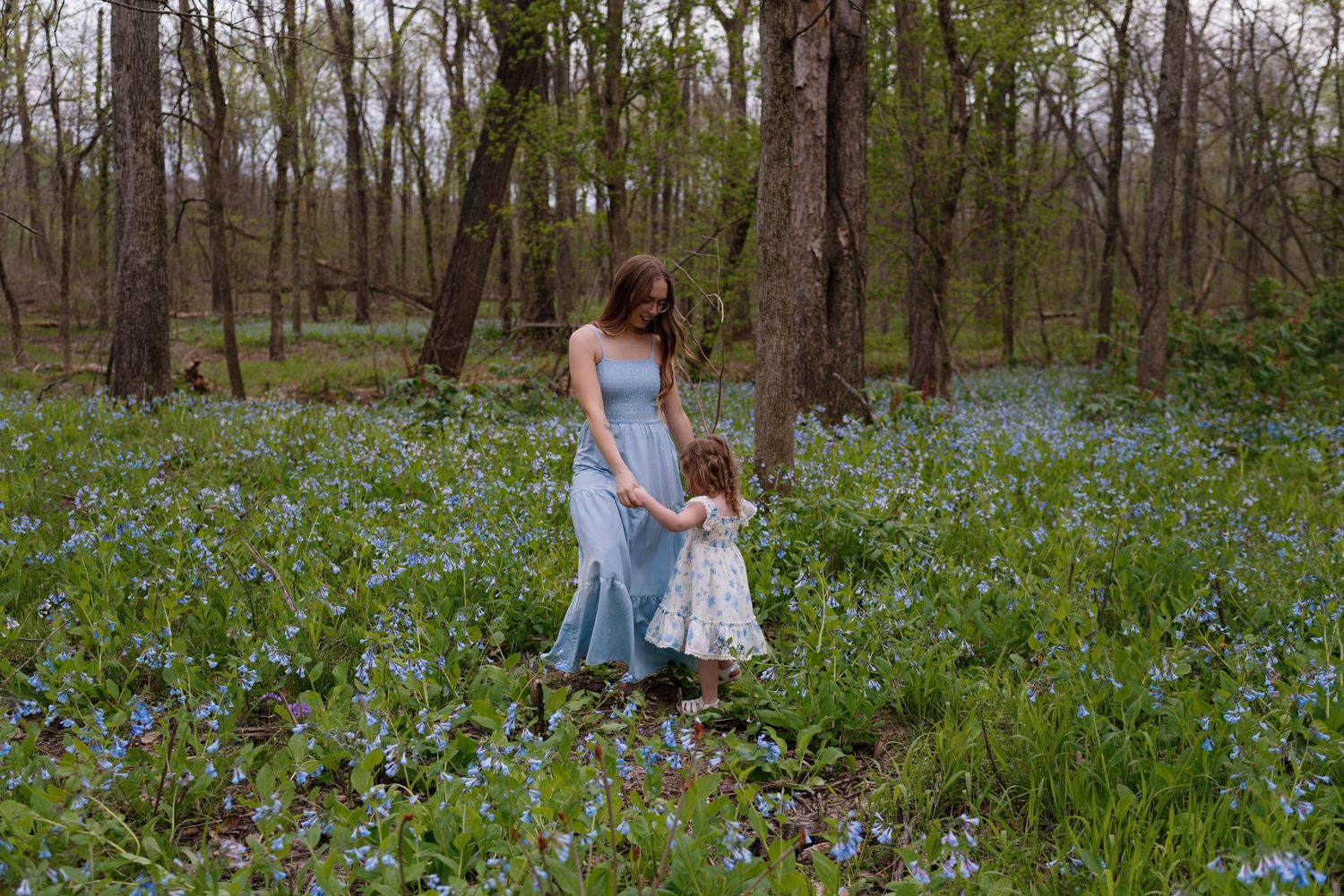 Mother and daughter in light blue dresses walk through a springtime forest filled with blooming bluebells.