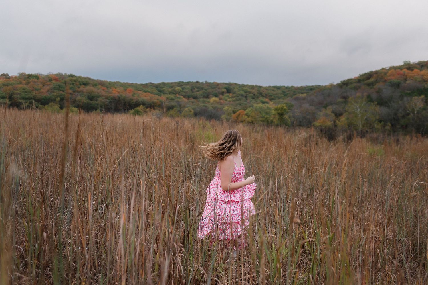 A young girl in a pink dress walks through tall golden prairie grass with autumn hills in the background.