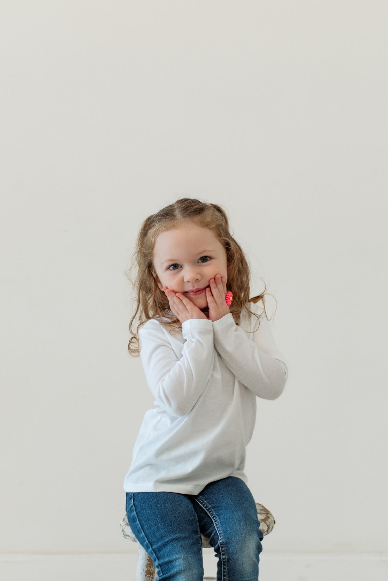 A young child in jeans and a white sweater sits playfully against a white background.