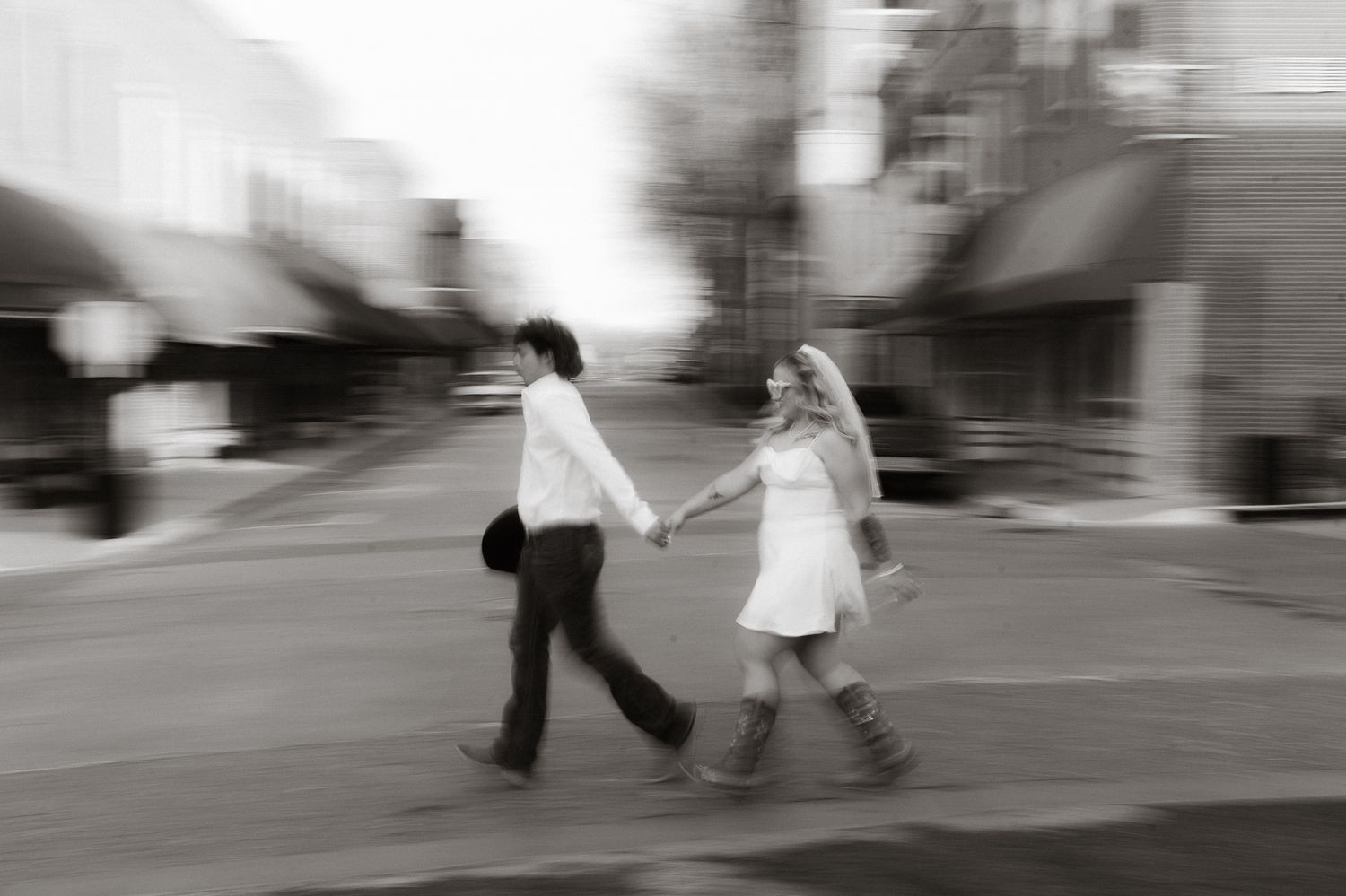 Black and white blurred photo of a couple holding hands and walking across a street in a town.