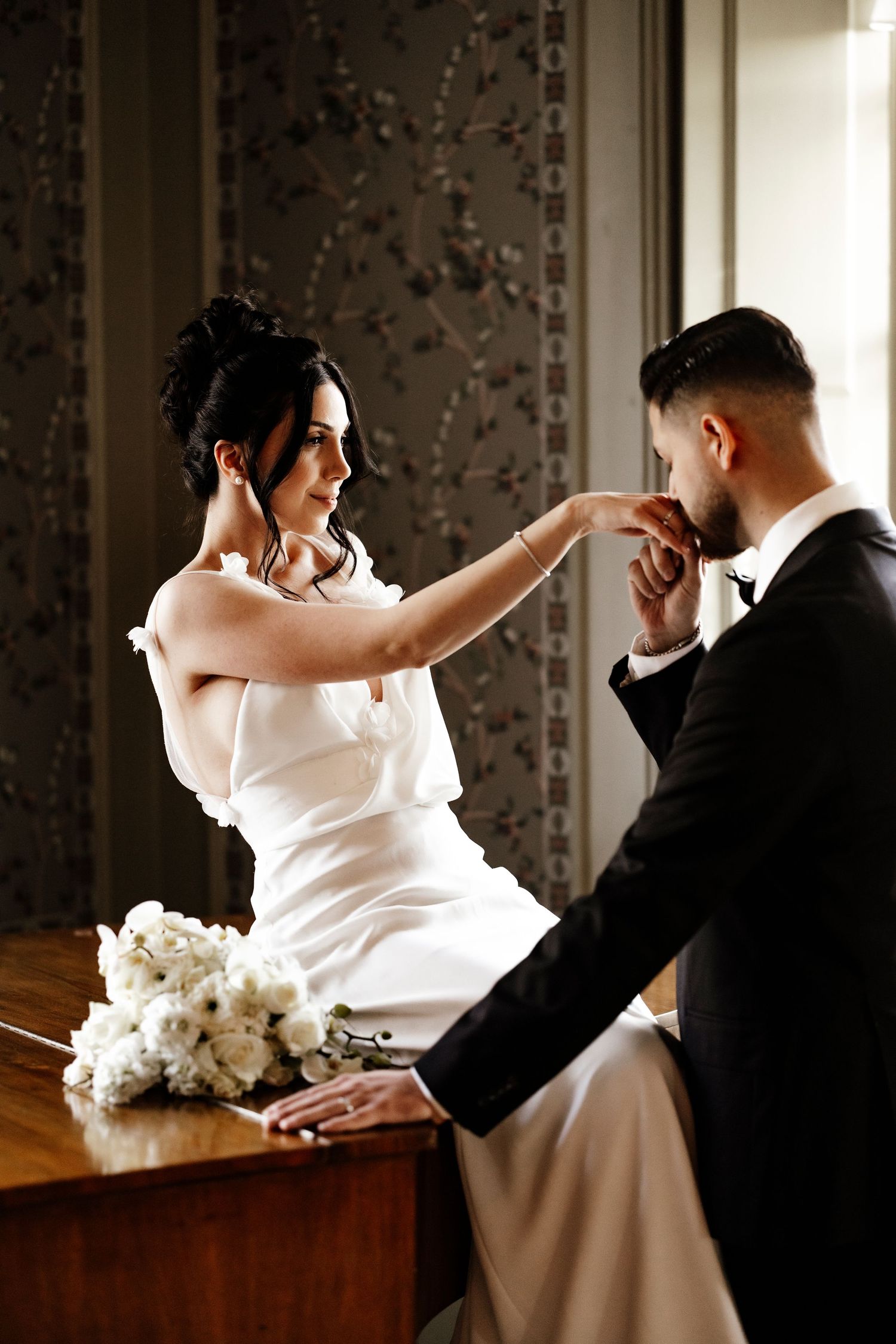 A romantic wedding moment captured as a bride in white and groom in black share an intimate gesture near a piano.
