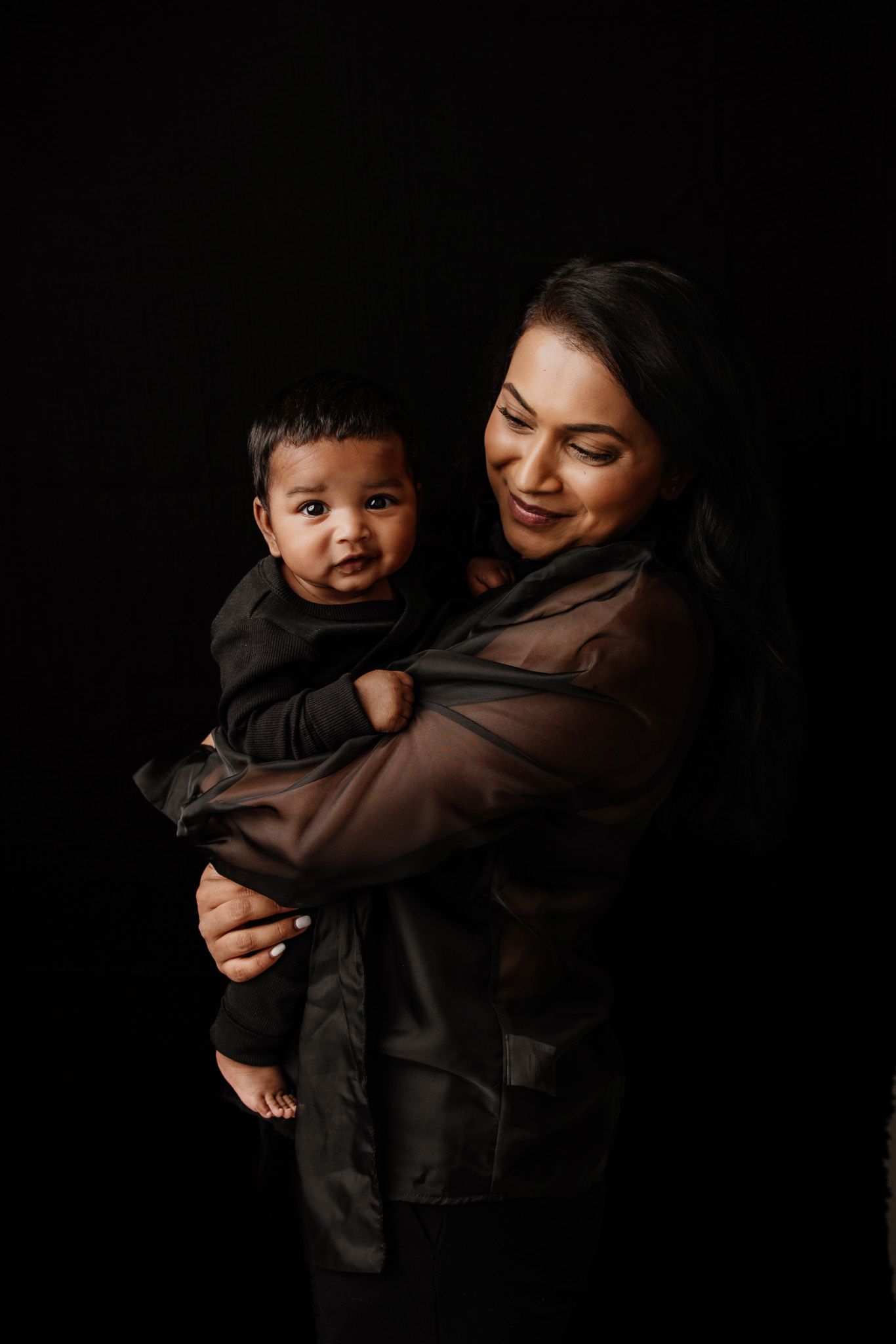 A woman in a black sheer jacket lovingly holds her baby, both dressed in black against a dark studio background.
