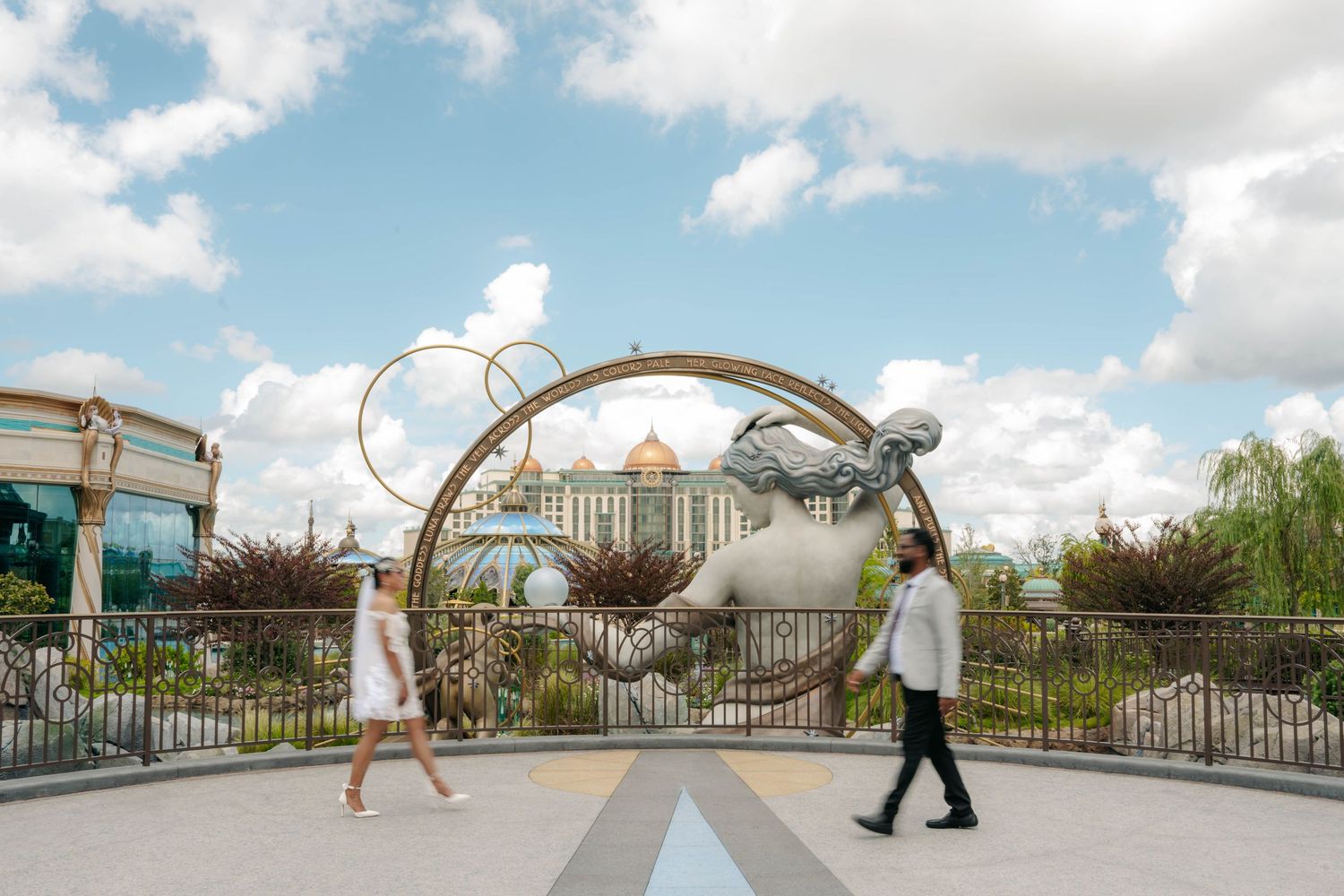 Two people walk past a grand entrance gate featuring a large circular sculpture at a modern theme park.