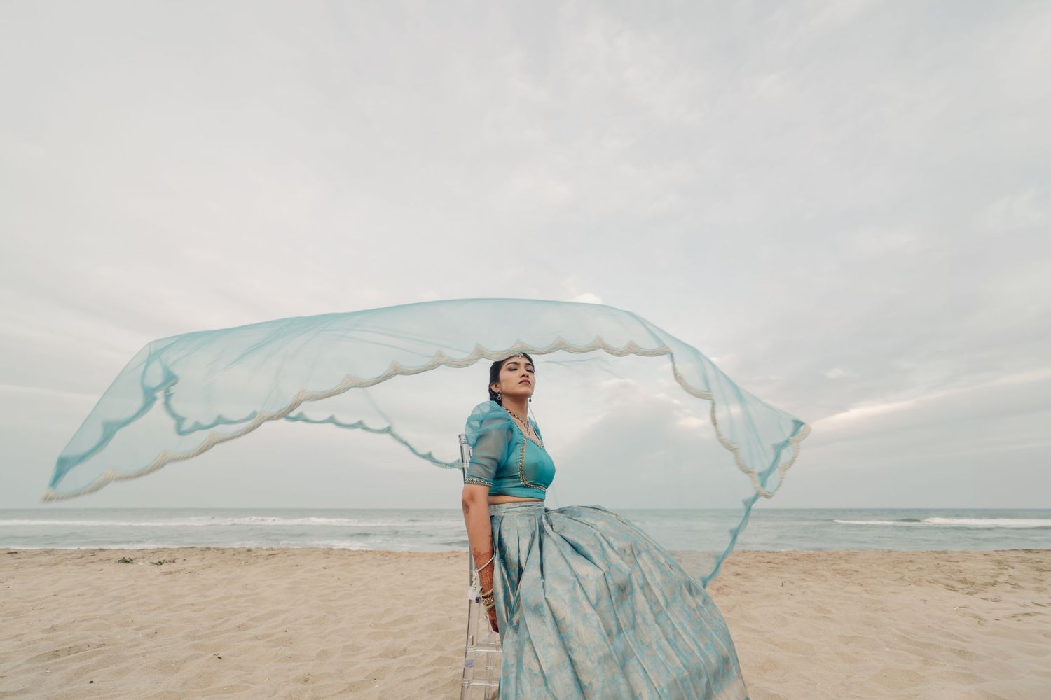 A flowing blue dress billows dramatically in the wind against a cloudy beach landscape.