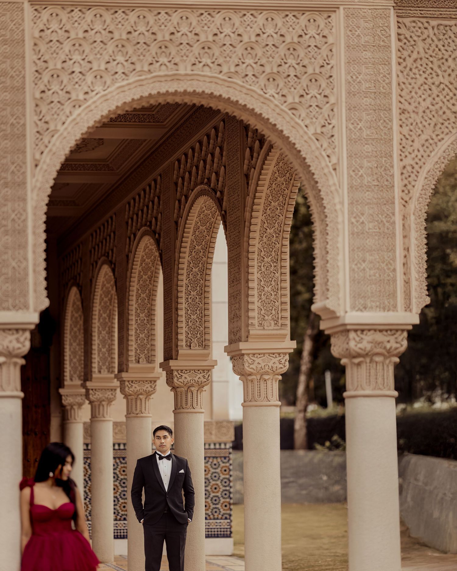 A couple poses in an ornate Moorish-style courtyard with intricately carved stone columns and arches.