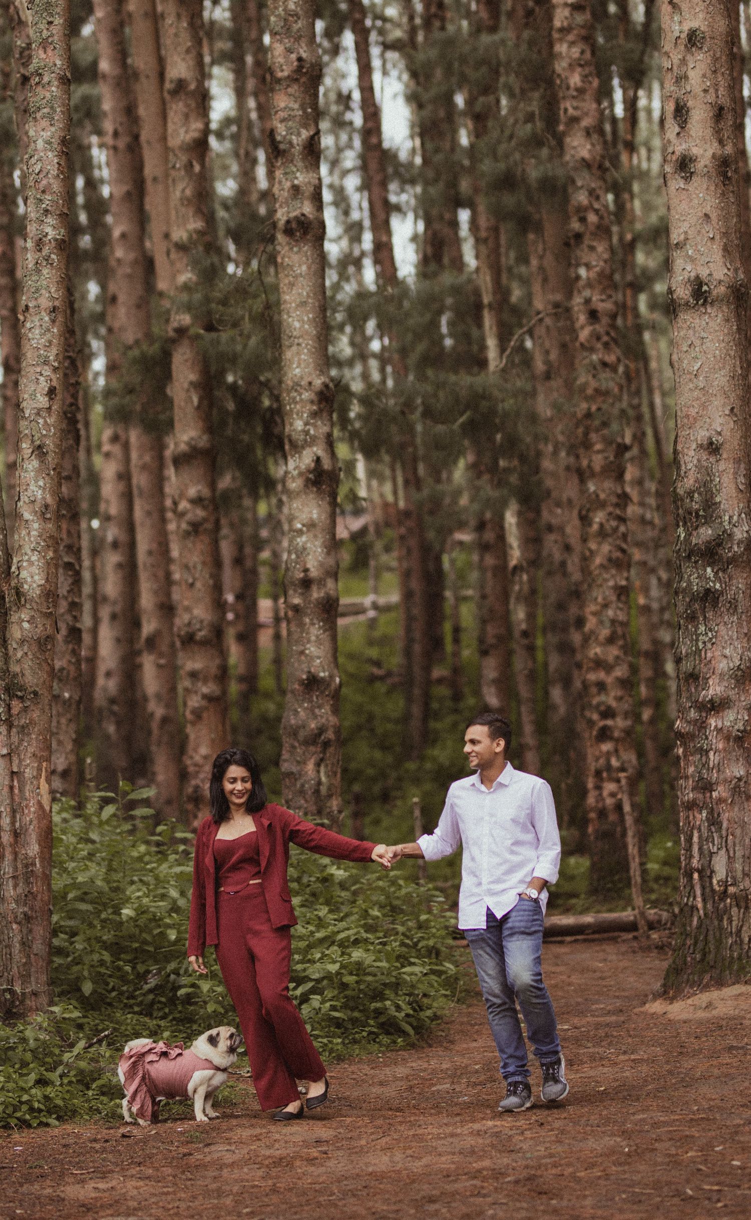 A couple playfully interacts while walking through a serene pine forest path on an autumn day.