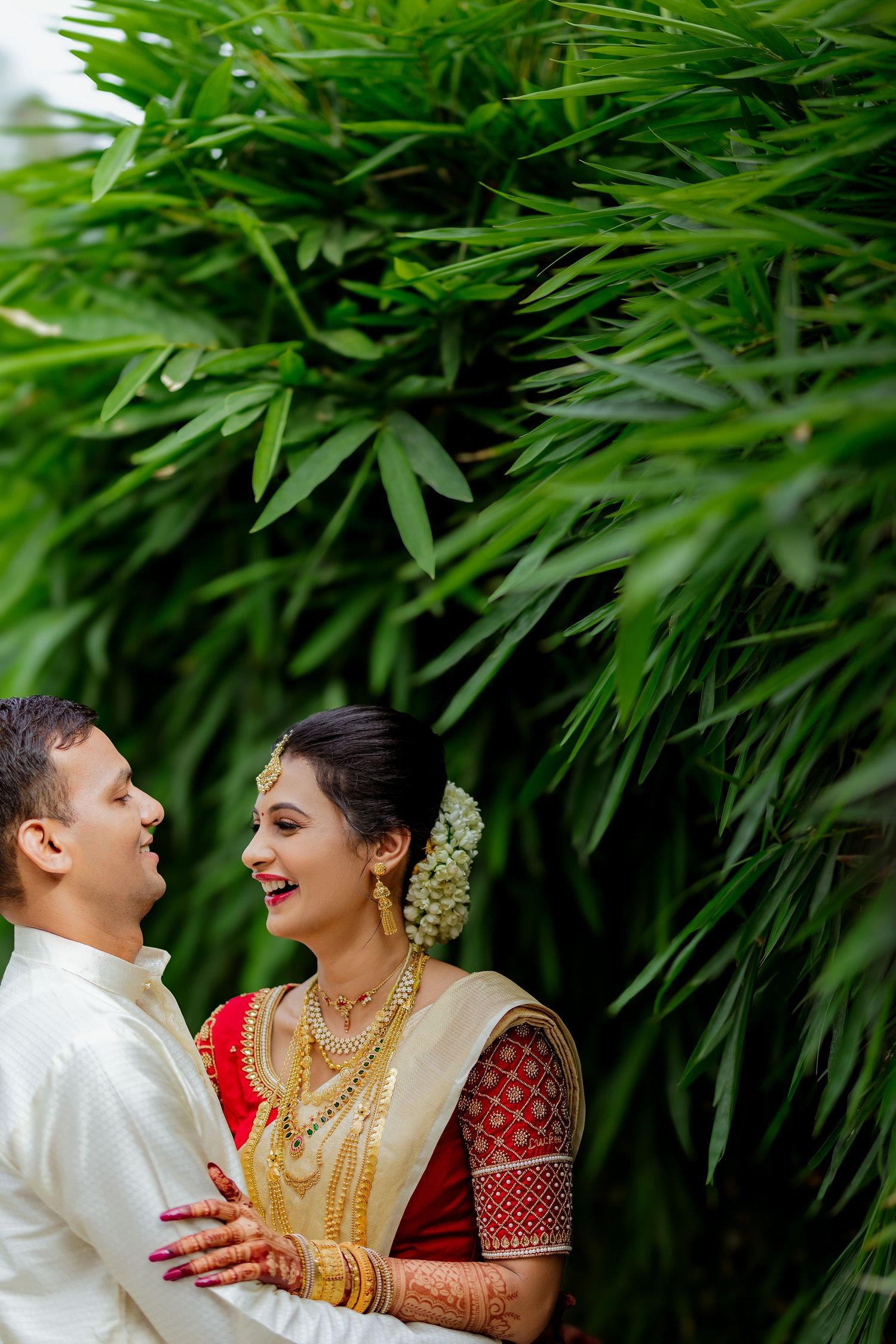 Traditional South Indian couple in wedding attire share a joyful moment against lush bamboo foliage background.