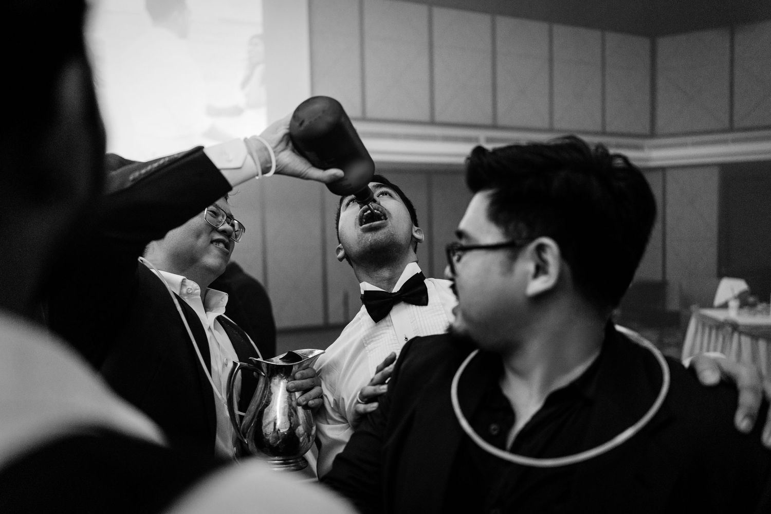 Black and white photo of men in formal attire at a wedding reception, one tilting head back drinking.