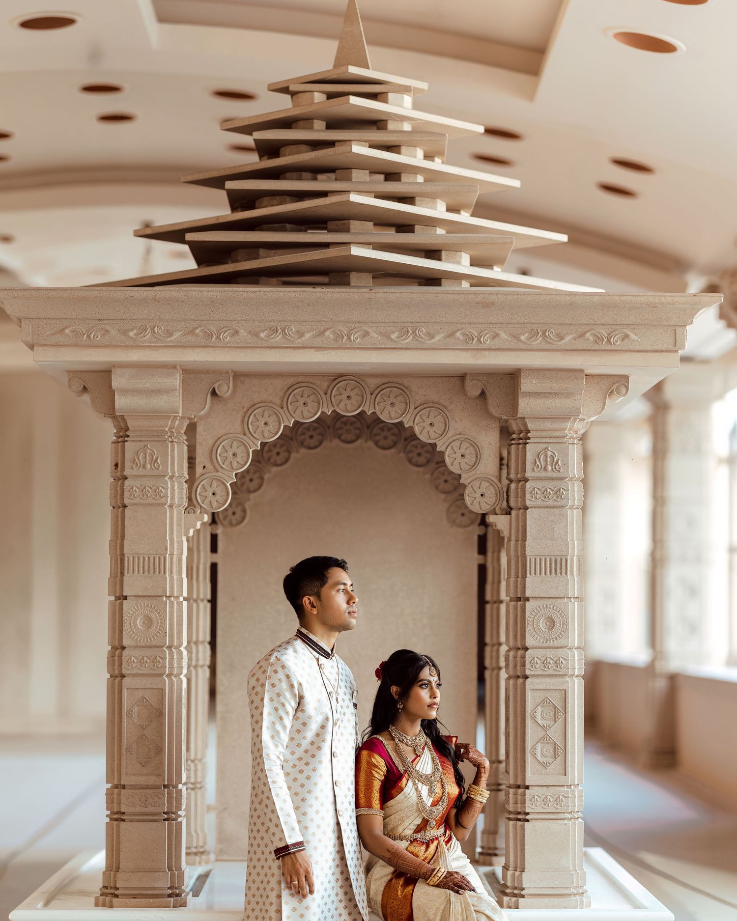 Indian couple in traditional attire posing inside an ornate white temple archway with decorative pillars.