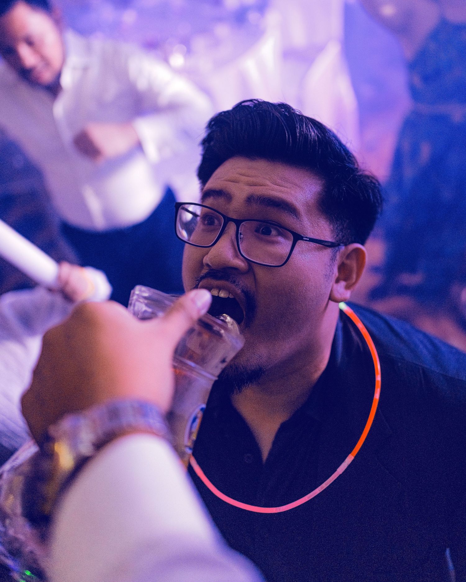 Man with glasses wearing a glow necklace drinks from a hookah pipe at a nightclub party.