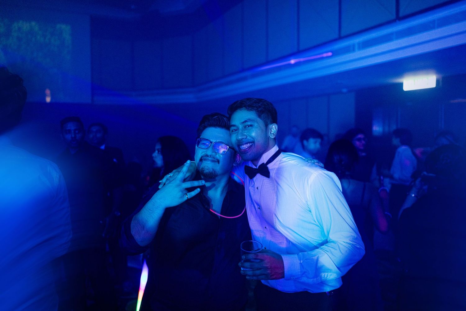 Two friends pose together on a dance floor under blue lights at a formal party event.