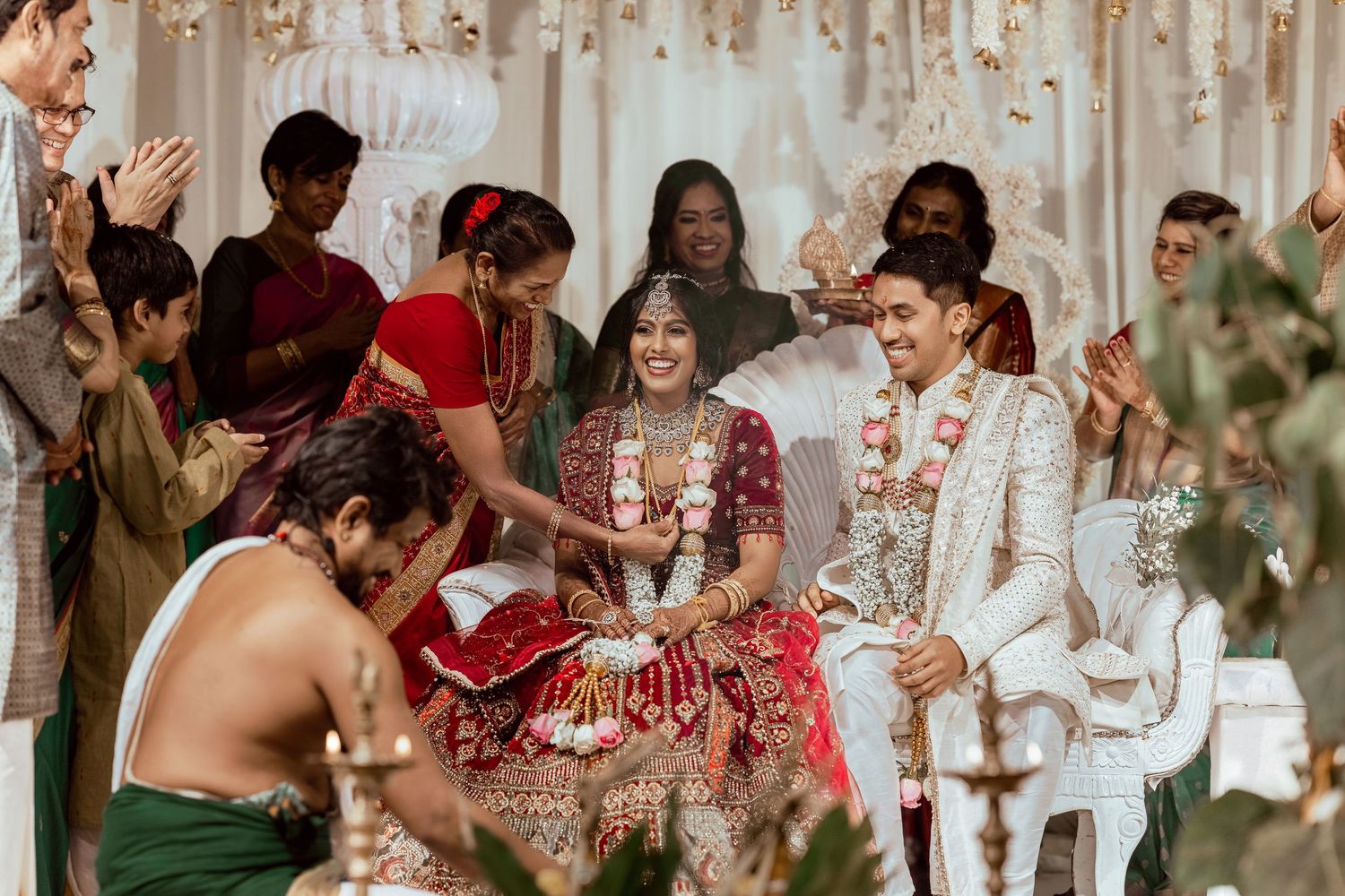 Joyful Indian wedding ceremony with bride in red lehenga and groom in white sherwani surrounded by celebrating guests.