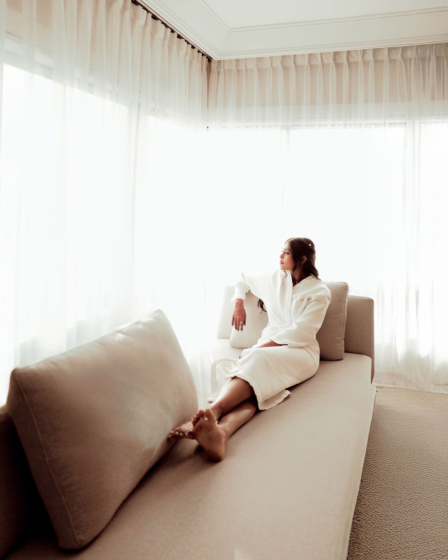 Woman in white robe relaxing on a beige sofa by sheer curtained windows in a bright, serene hotel room.