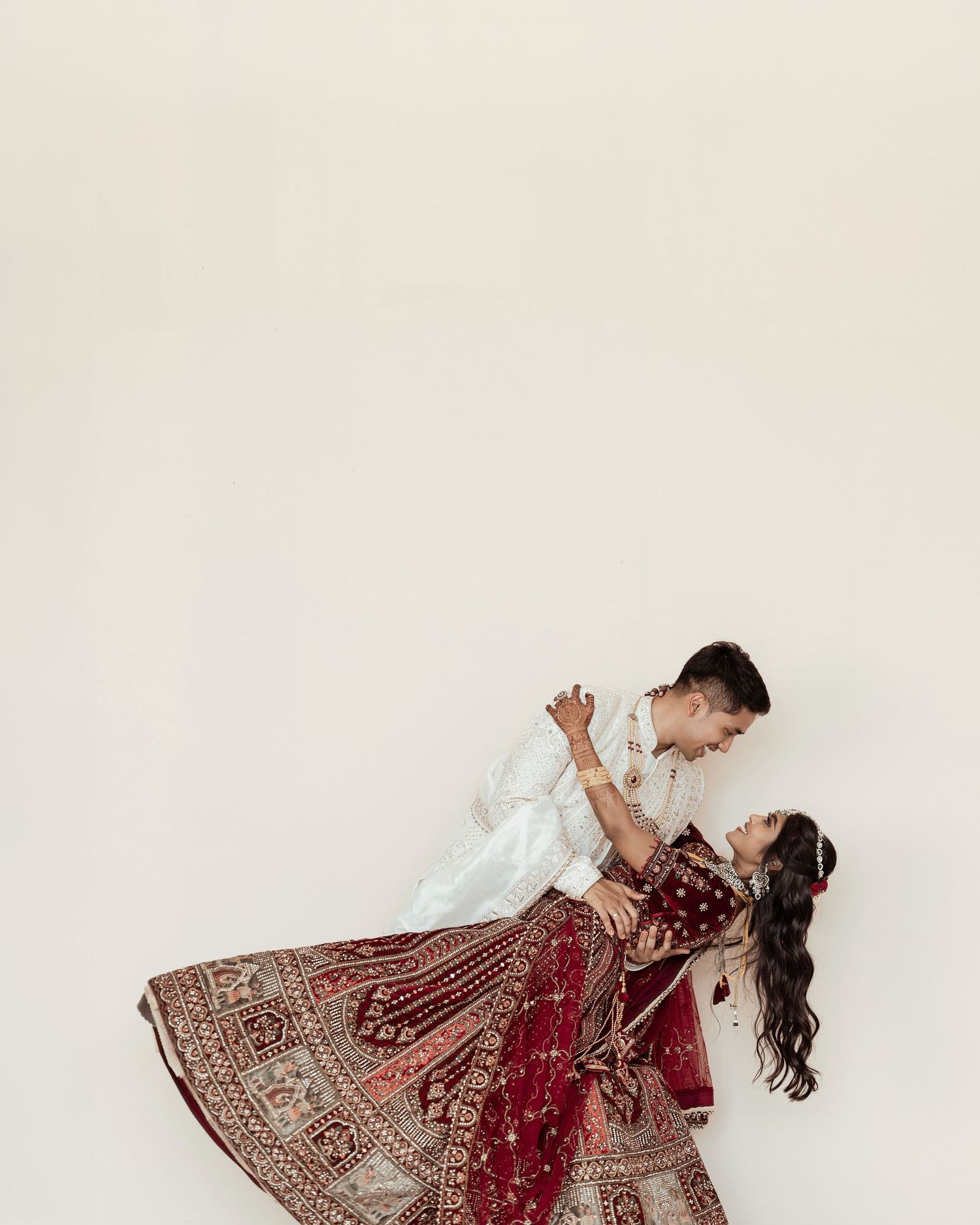 Couple in traditional Indian wedding attire dancing, bride in red embroidered lehenga, groom in white kurta, against white backdrop.