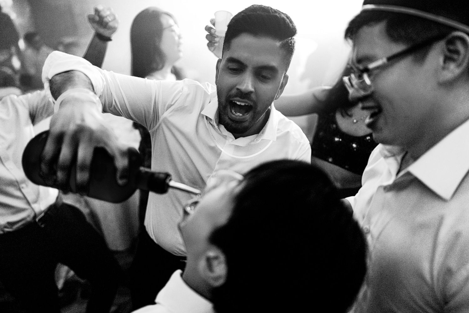 Black and white photo of men at a party, one pouring a drink into another's mouth, laughing and celebrating.