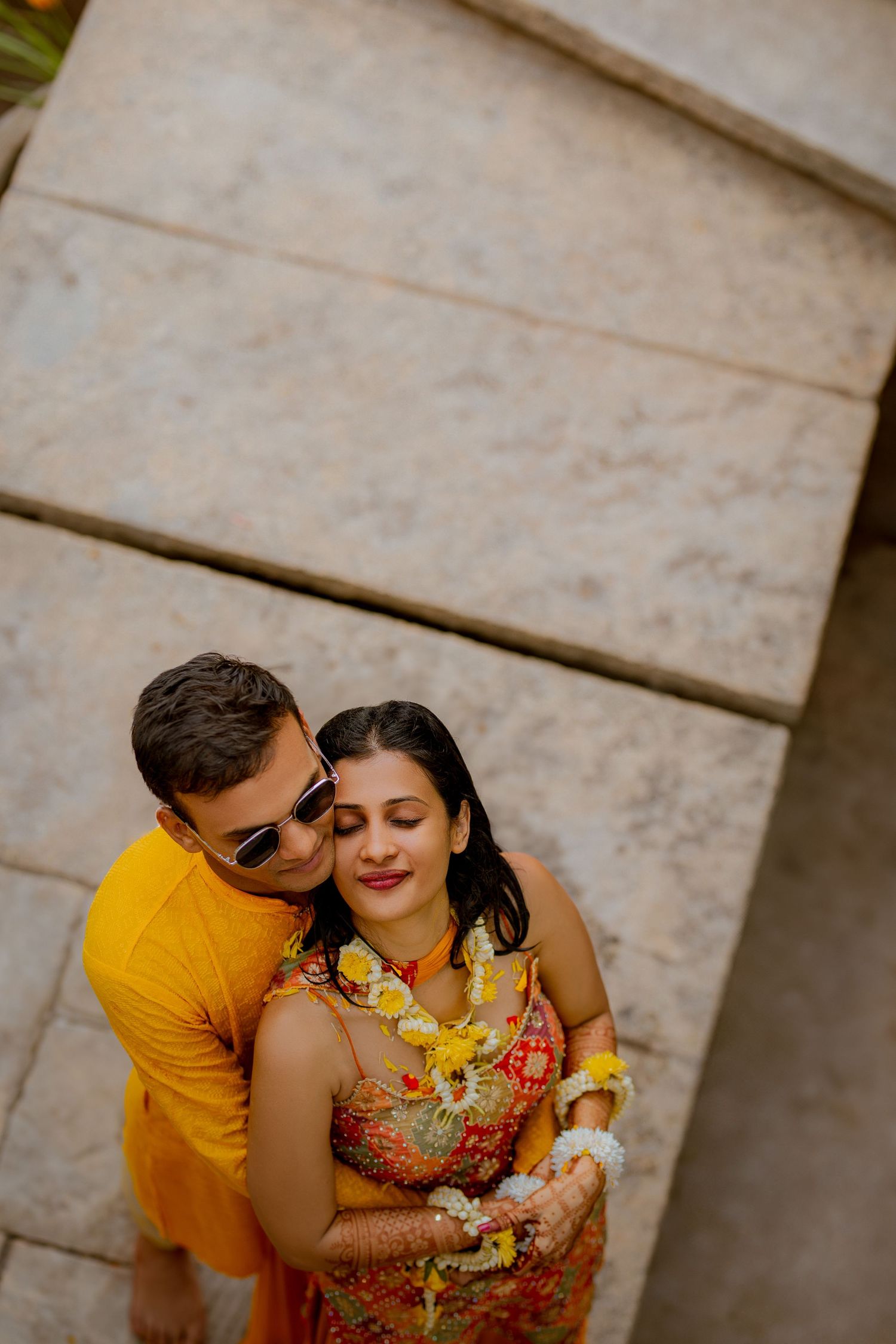 A couple embraces outdoors, with one wearing a yellow shirt and the other in a floral dress with a flower garland.