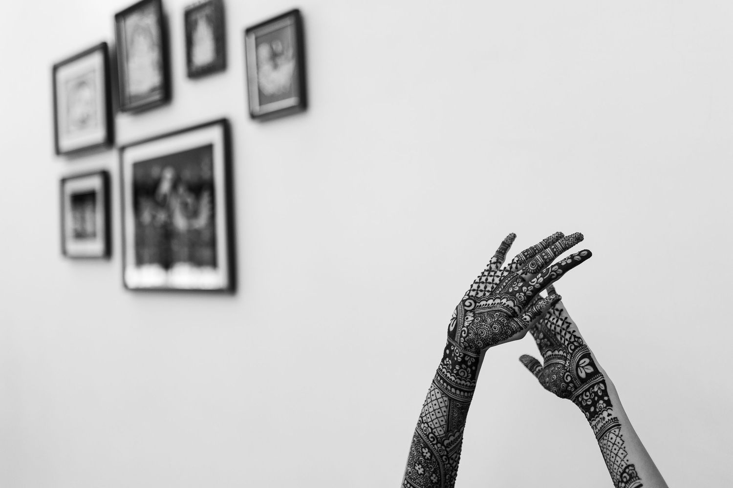 Black and white photo of hands decorated with intricate henna designs against a wall with framed artwork.