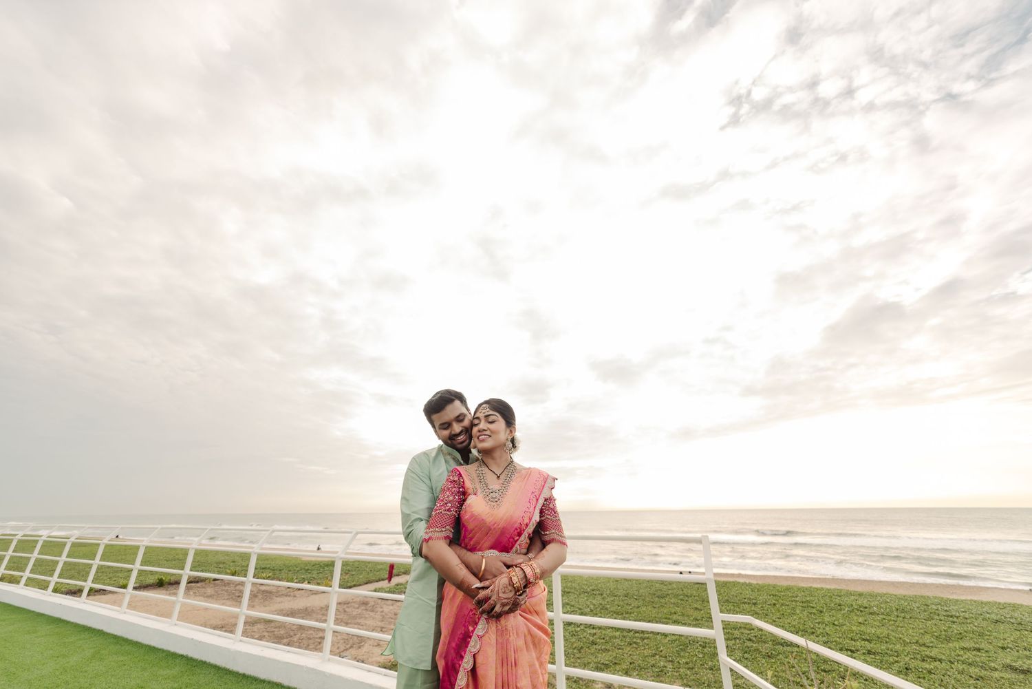 Romantic couple poses against sunset sky at a horse racing track with white fencing.