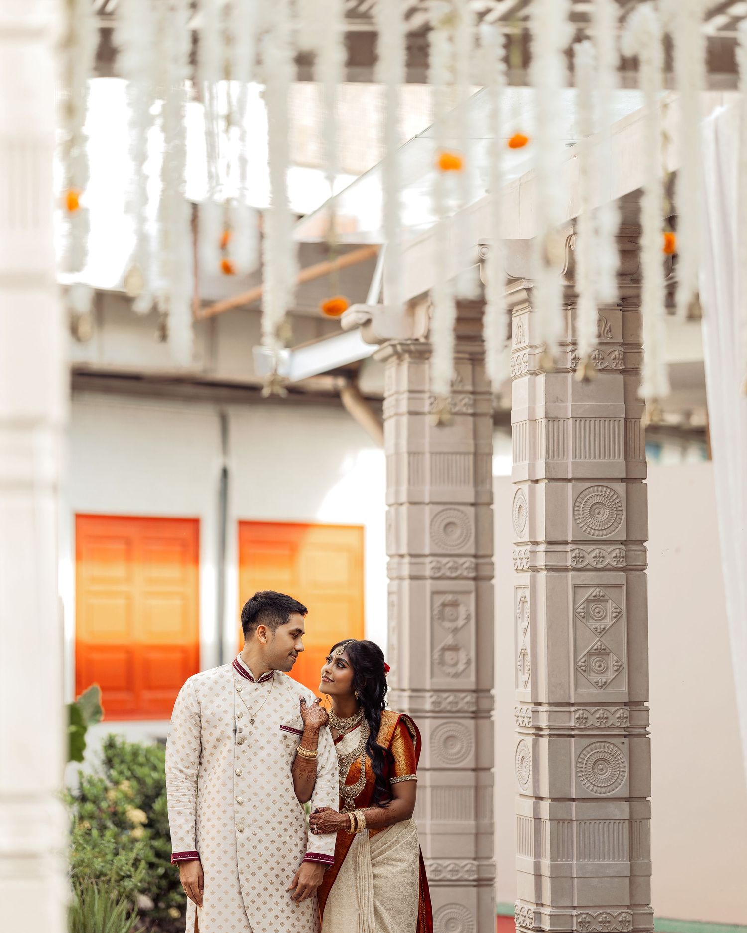 Indian couple in traditional wedding attire posing beside ornate white temple pillars with floral decorations.
