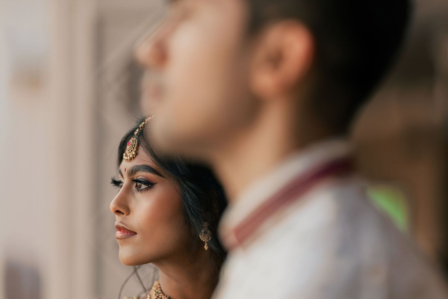 Indian bride in gold jewelry gazes softly, with groom in white sherwani blurred in foreground.