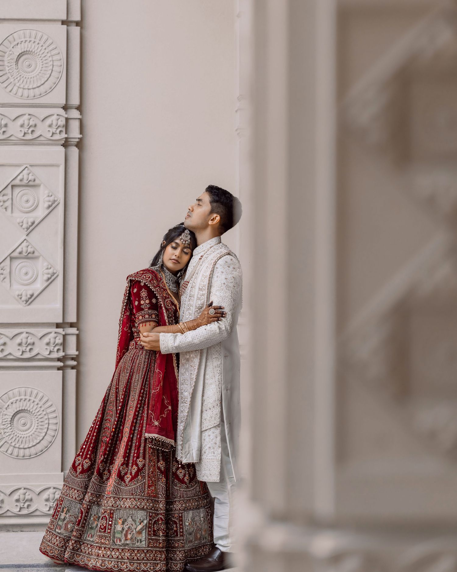 Indian bride in red lehenga and groom in white sherwani embrace in an elegant wedding portrait.