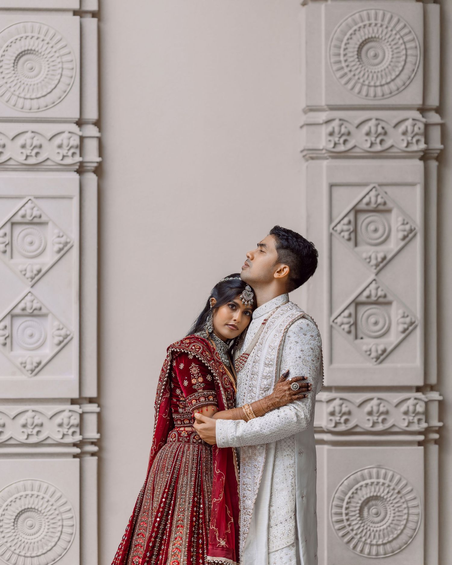 Couple in traditional South Asian wedding attire embracing before ornate white carved wall, bride in red lehenga.