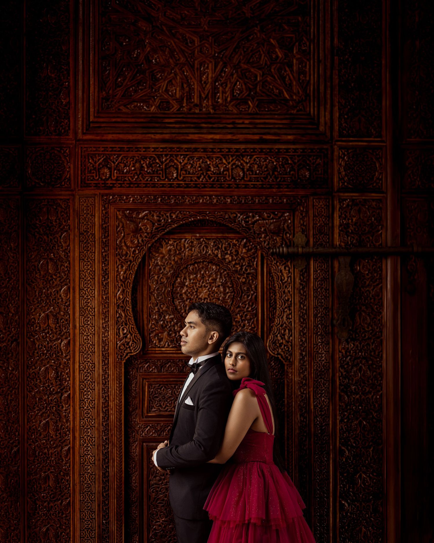 Couple in formal attire posing against an ornate carved wooden door, man in tuxedo and woman in red gown.