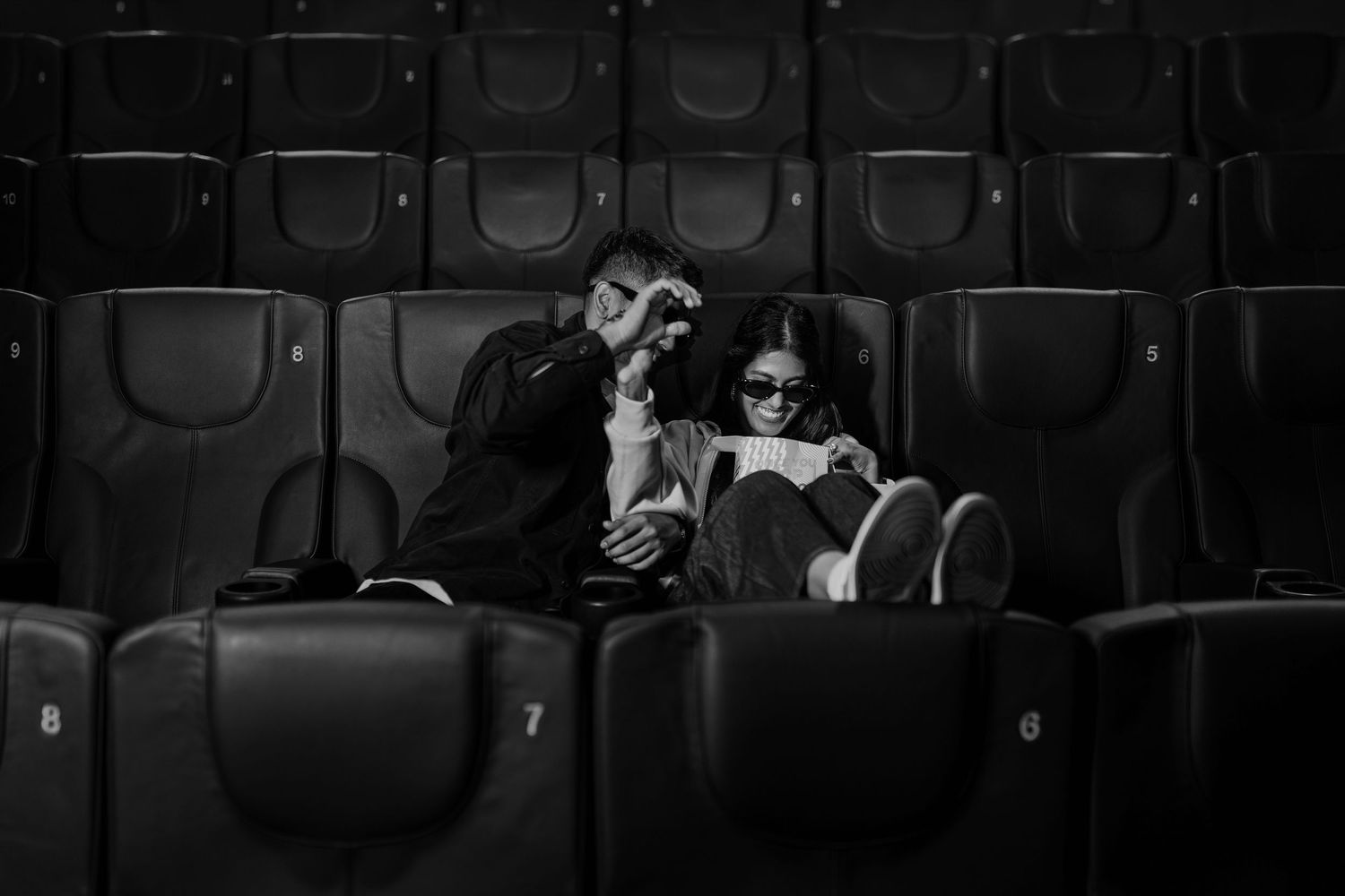 Black and white photo of a couple sitting close together in empty theater seats, sharing an intimate moment.