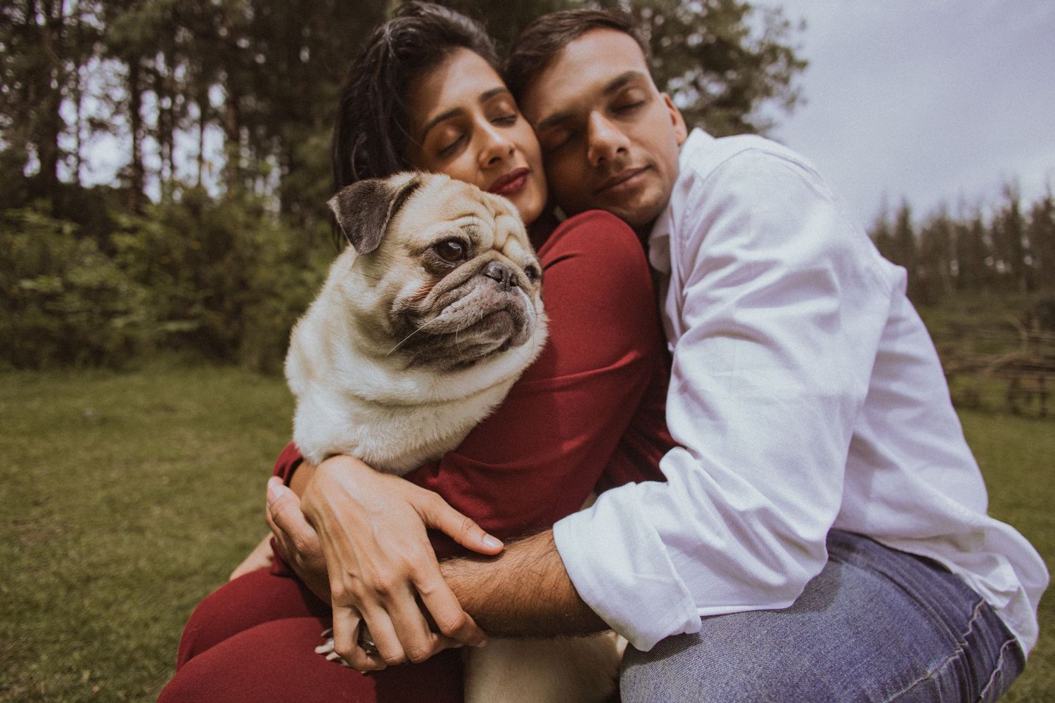 A couple in burgundy and white outfits poses together in a natural outdoor setting with their French Bulldog.